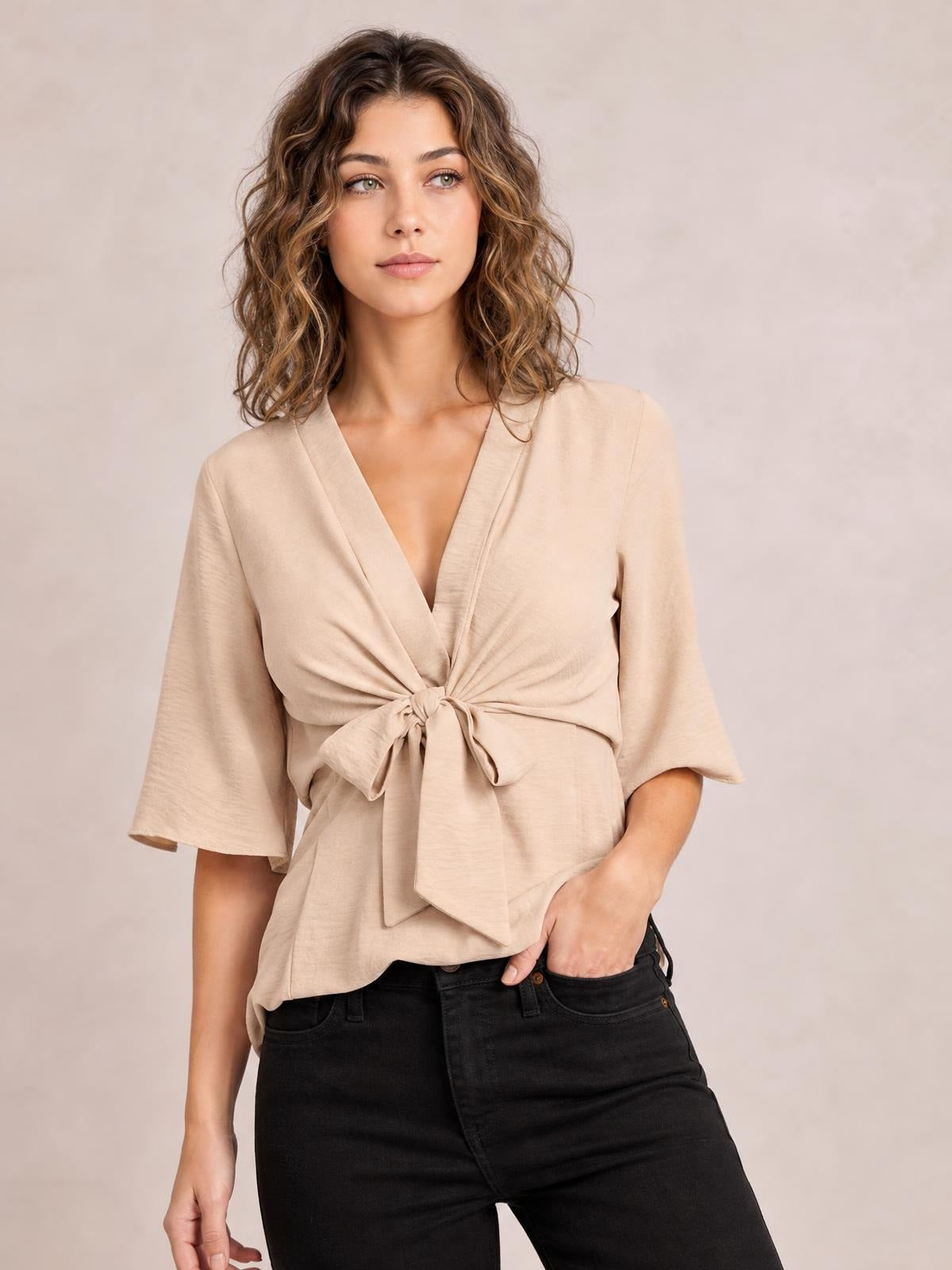 Model wears a solid beige V-neck tie-front blouse with twisted detailing, three-quarter sleeves, and a relaxed long silhouette.