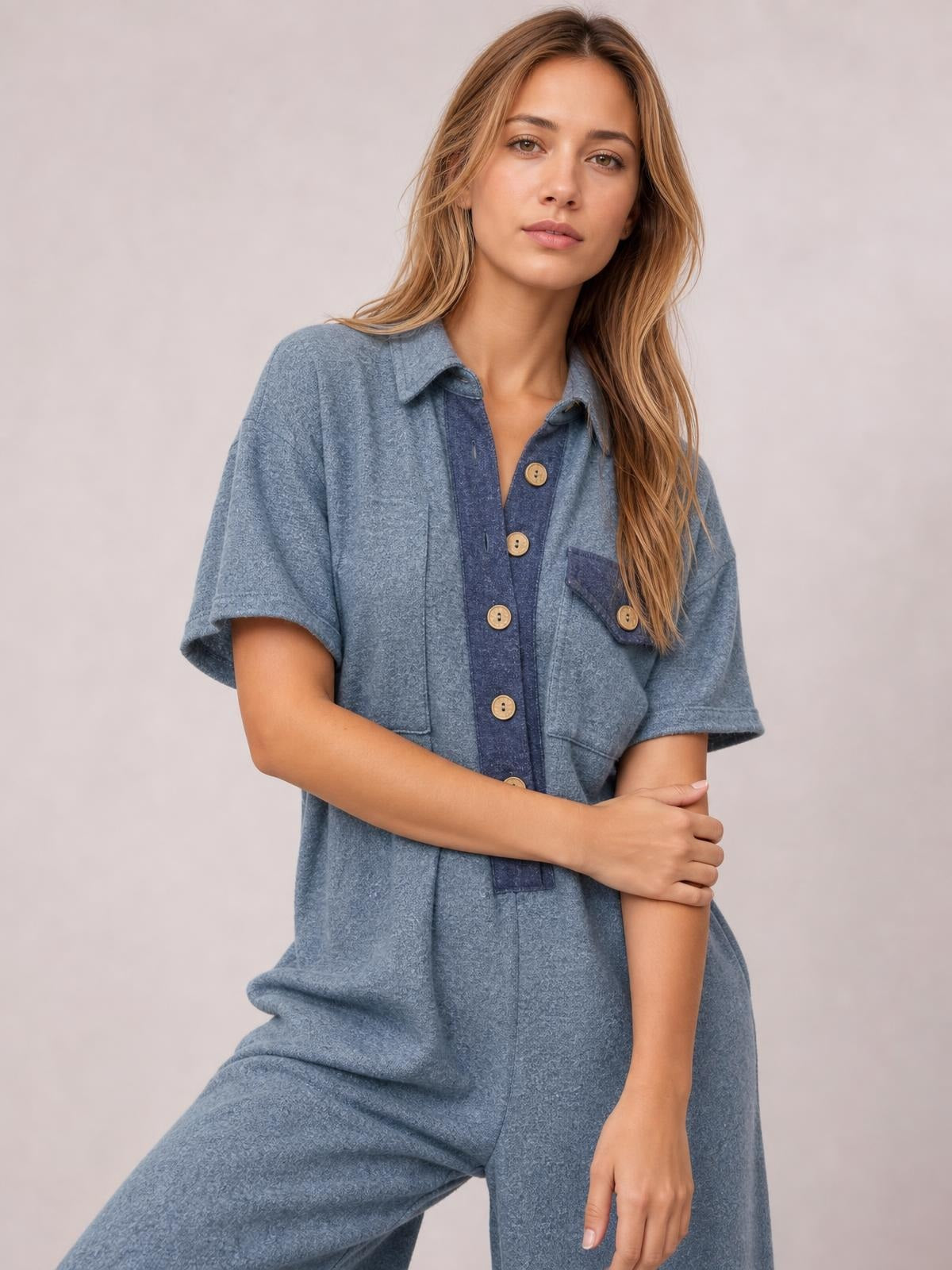 Model poses in a blue short-sleeve wide-leg jumpsuit with a buttoned placket, pointed collar, and two visible chest pockets.