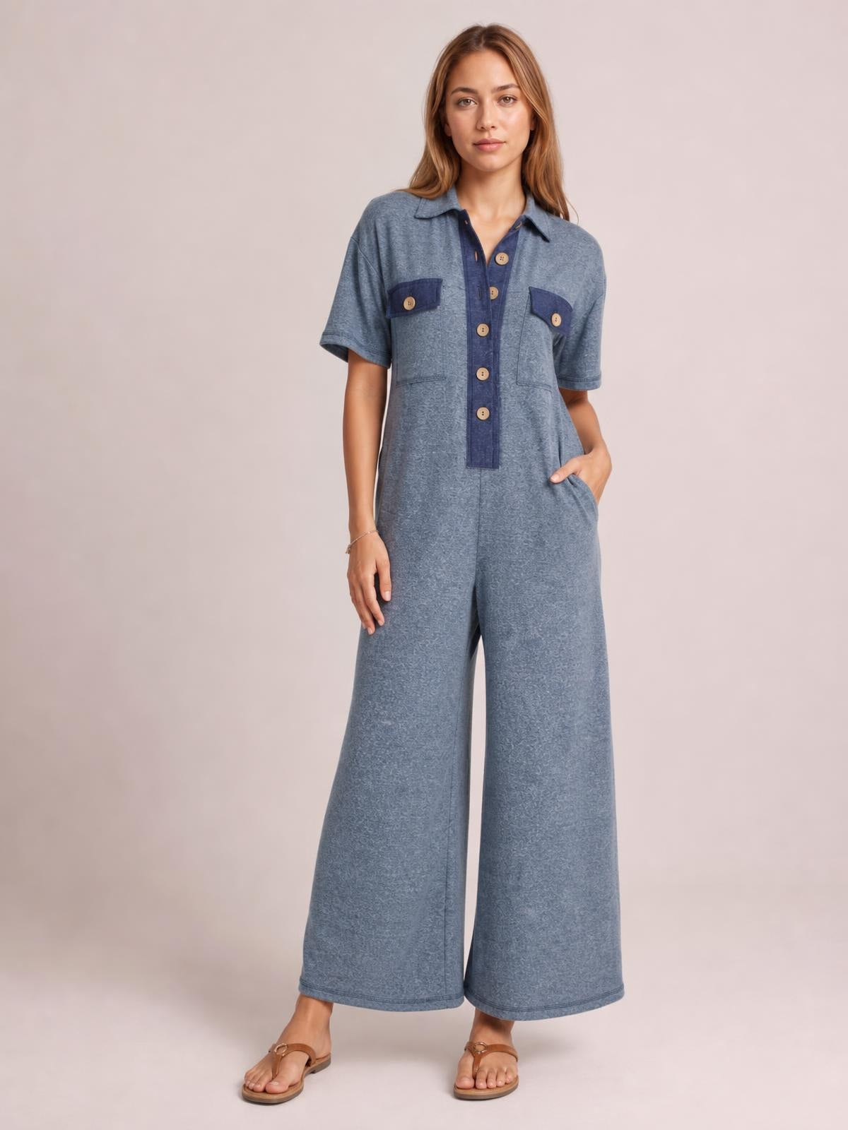 A model wears a blue short-sleeve jumpsuit with a buttoned placket, chest pockets, and a relaxed wide-leg silhouette, posing with one hand in a side pocket.