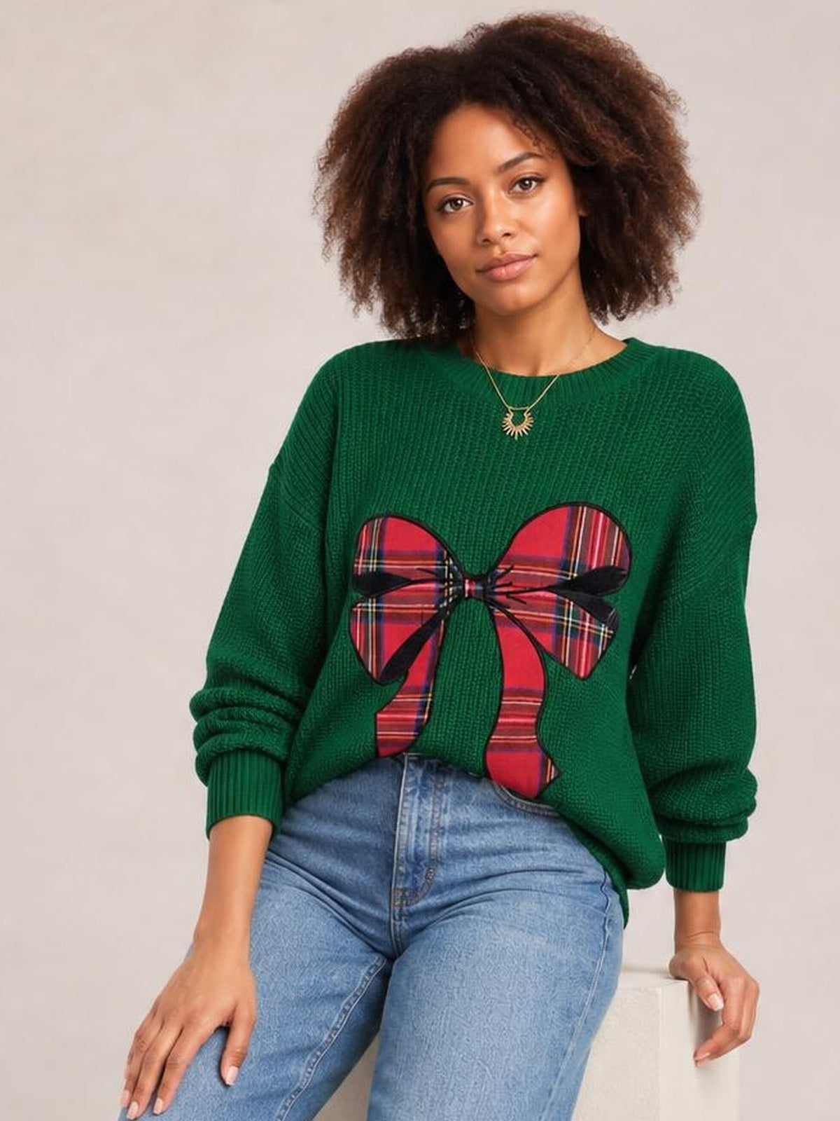 A seated model wears a green Christmas sweater with a round neck, long sleeves, ribbed cuffs, and a large red plaid bow across the front.