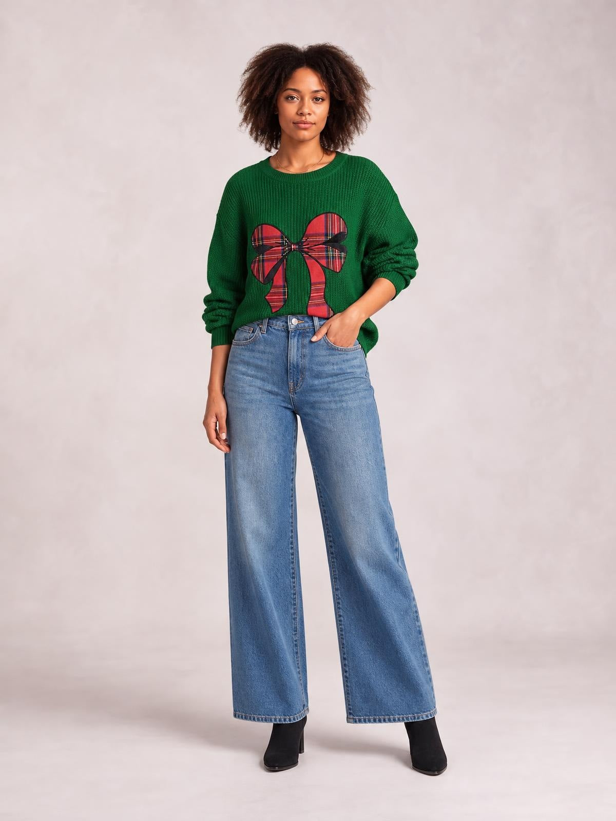 Model wears a green round-neck Christmas sweater with a large red plaid bow, ribbed cuffs, relaxed silhouette, and one hand in her jeans pocket.