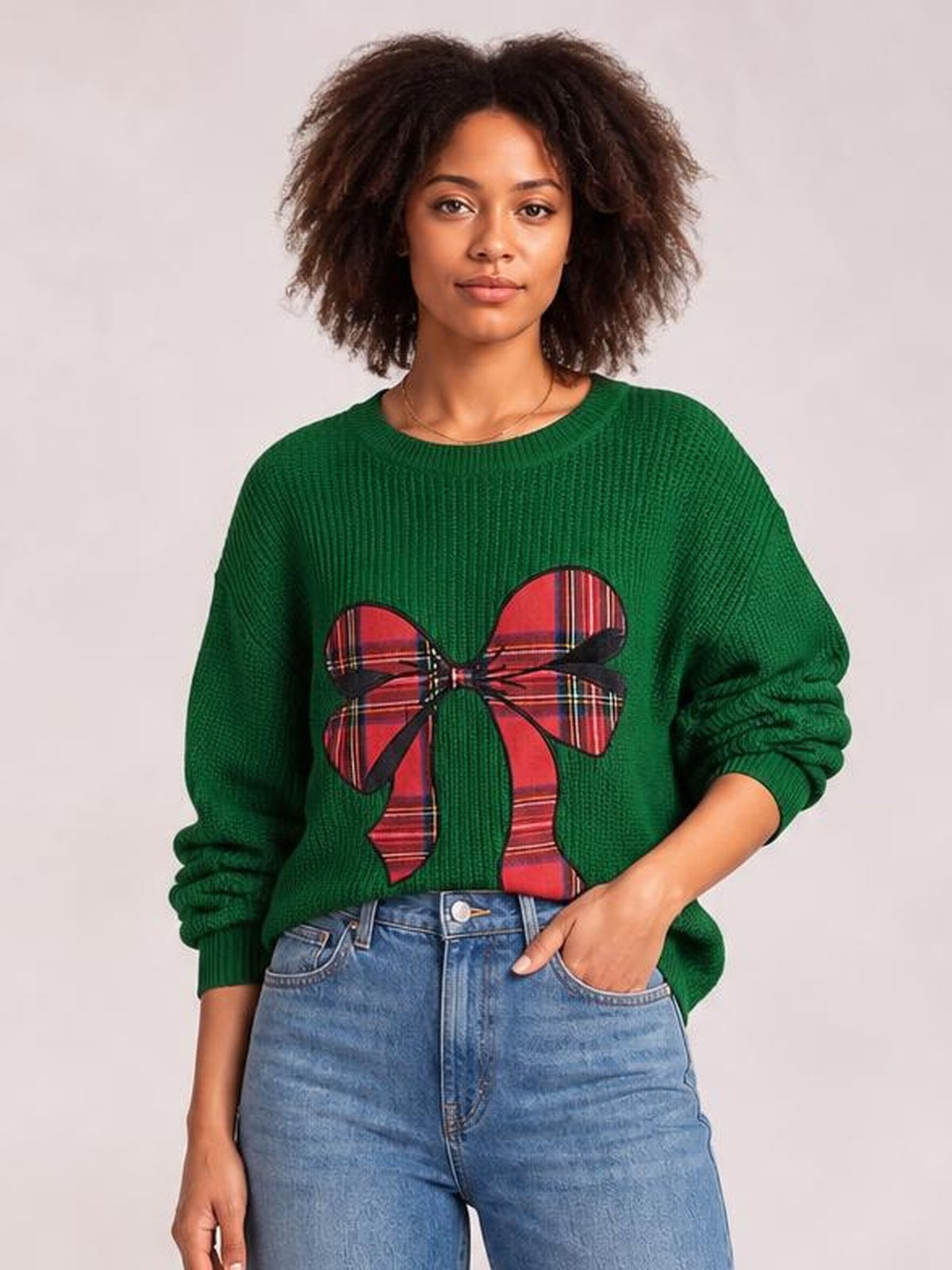 Model poses with one hand in her jeans pocket wearing a green round-neck Christmas sweater with long sleeves and a large red plaid bow graphic.
