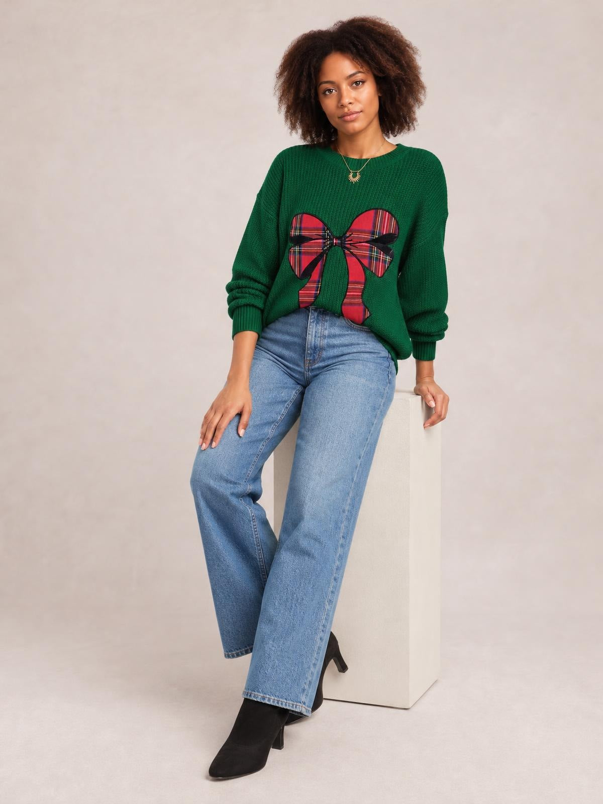 Model seated in a green round-neck Christmas sweater with a large red plaid bow applique, relaxed long sleeves, ribbed cuffs, and a loose silhouette.