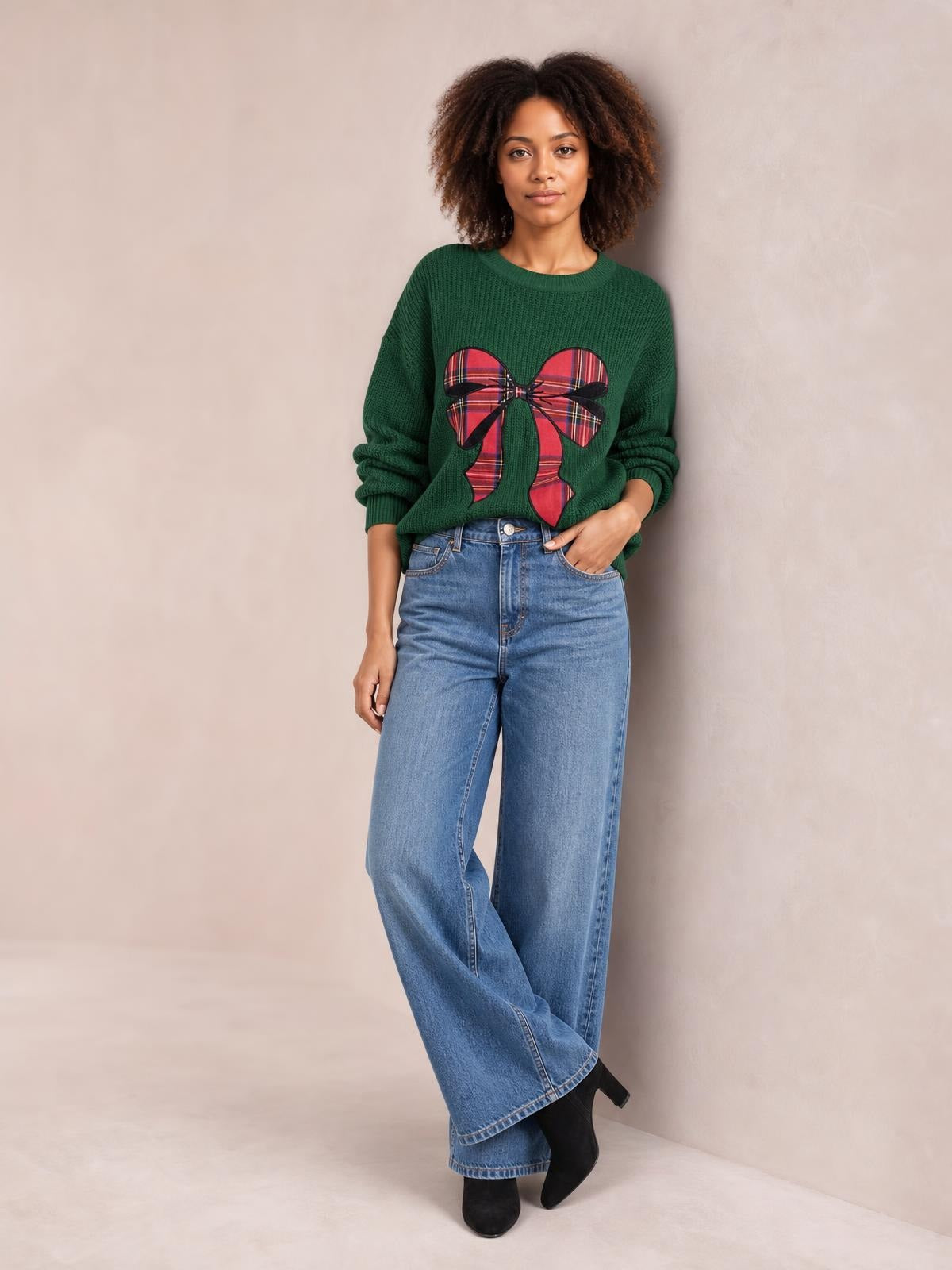 A model poses in a forest-green round-neck Christmas sweater with a red plaid bow design, ribbed cuffs, relaxed sleeves, and a slightly loose silhouette.