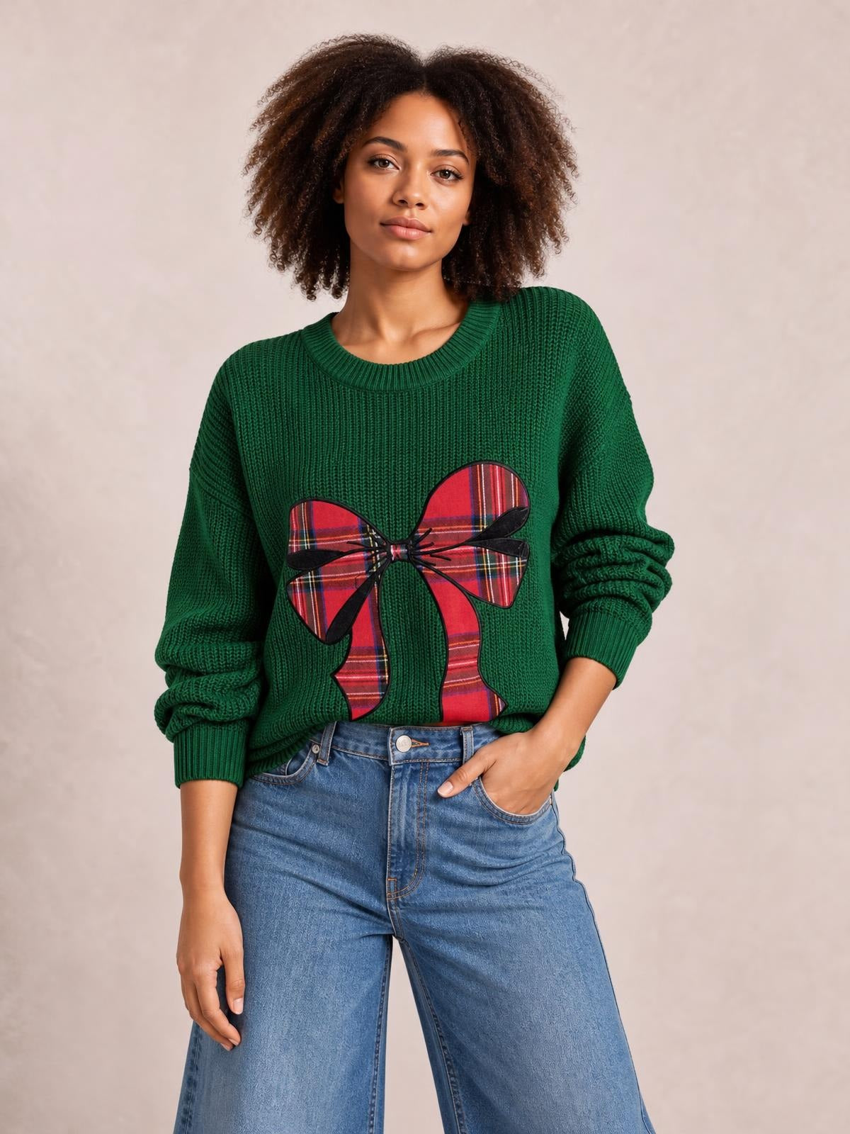 Model poses in a green round-neck Christmas sweater with ribbed cuffs and a large red plaid bow applique across the front.