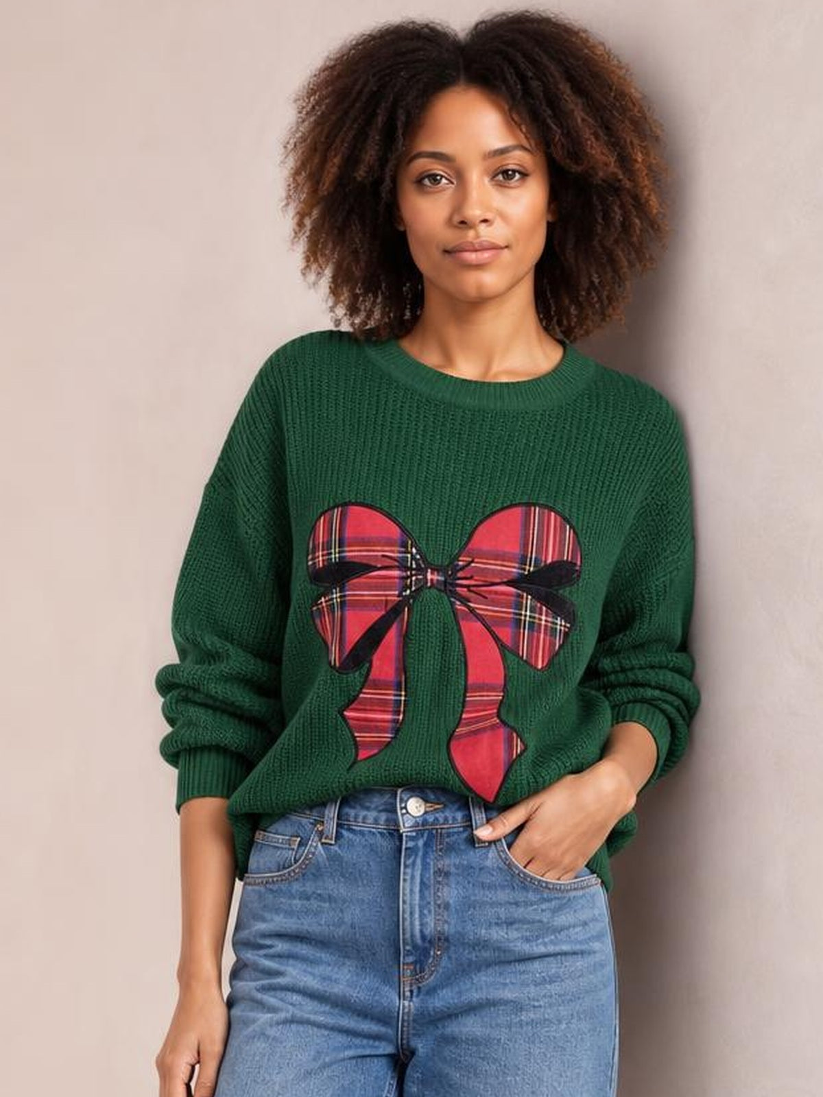A woman poses in a dark green round-neck Christmas sweater with a large red plaid bow design, ribbed cuffs and hem, and a relaxed silhouette.