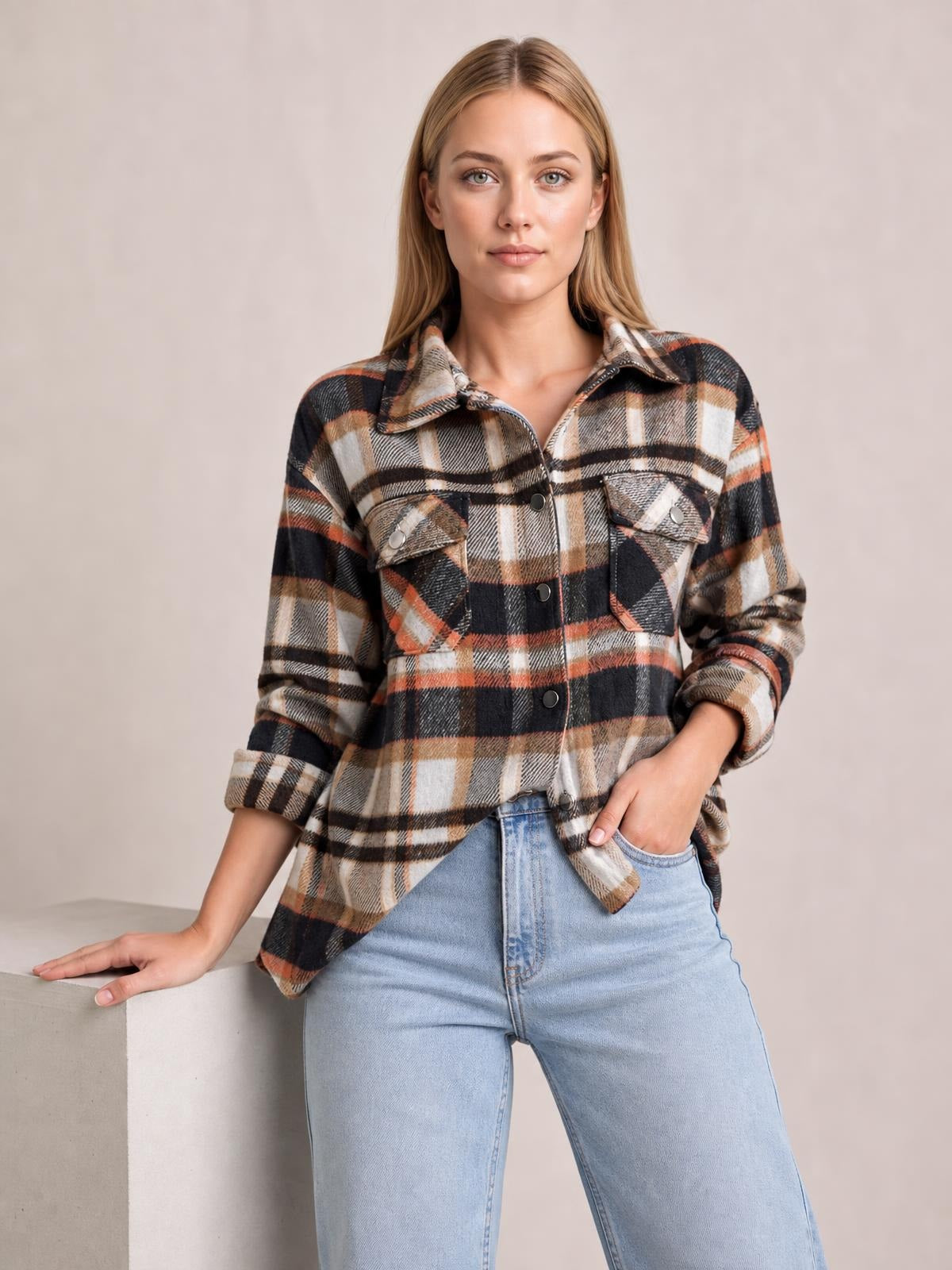 Model stands in a black, tan, cream, and orange plaid button-front shirt jacket with a collar, long sleeves, dropped shoulders, and two breast pockets, one hand in her jeans pocket.
