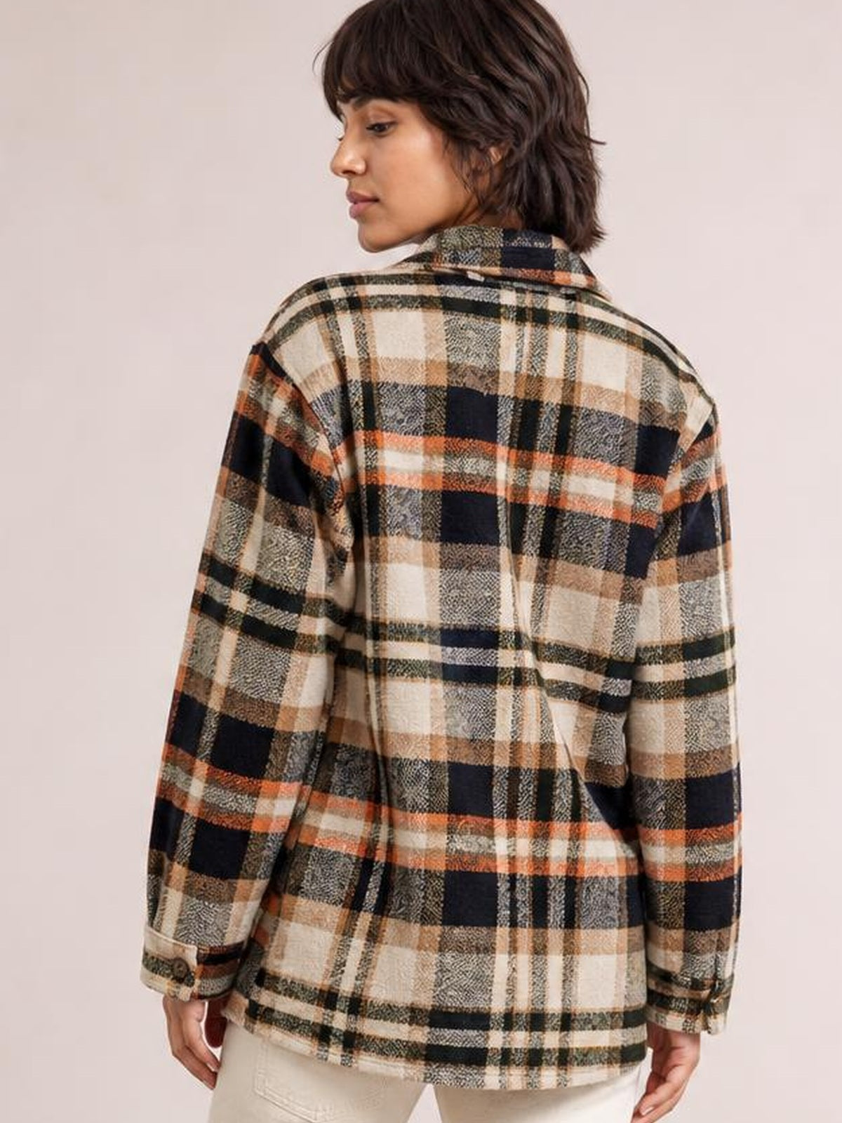 Model looks over her shoulder wearing a relaxed plaid button-front shirt jacket in cream, black, tan, and orange with a collared neckline and dropped shoulders.
