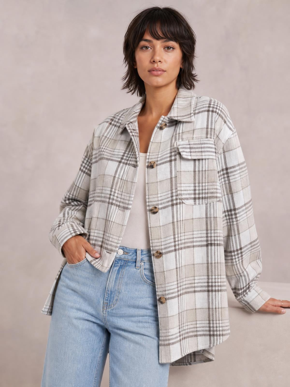 Model poses in a cream and gray plaid button-up jacket with a collared neckline, front buttons, flap pockets, relaxed silhouette, and rolled cuffs.