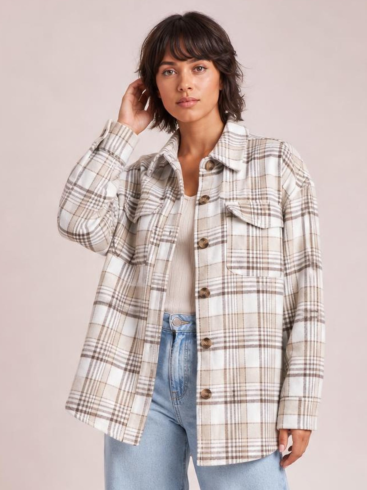 Model wears a cream and brown plaid button-up jacket with long sleeves, flap pockets, brown buttons, and a relaxed straight silhouette while touching her hair.
