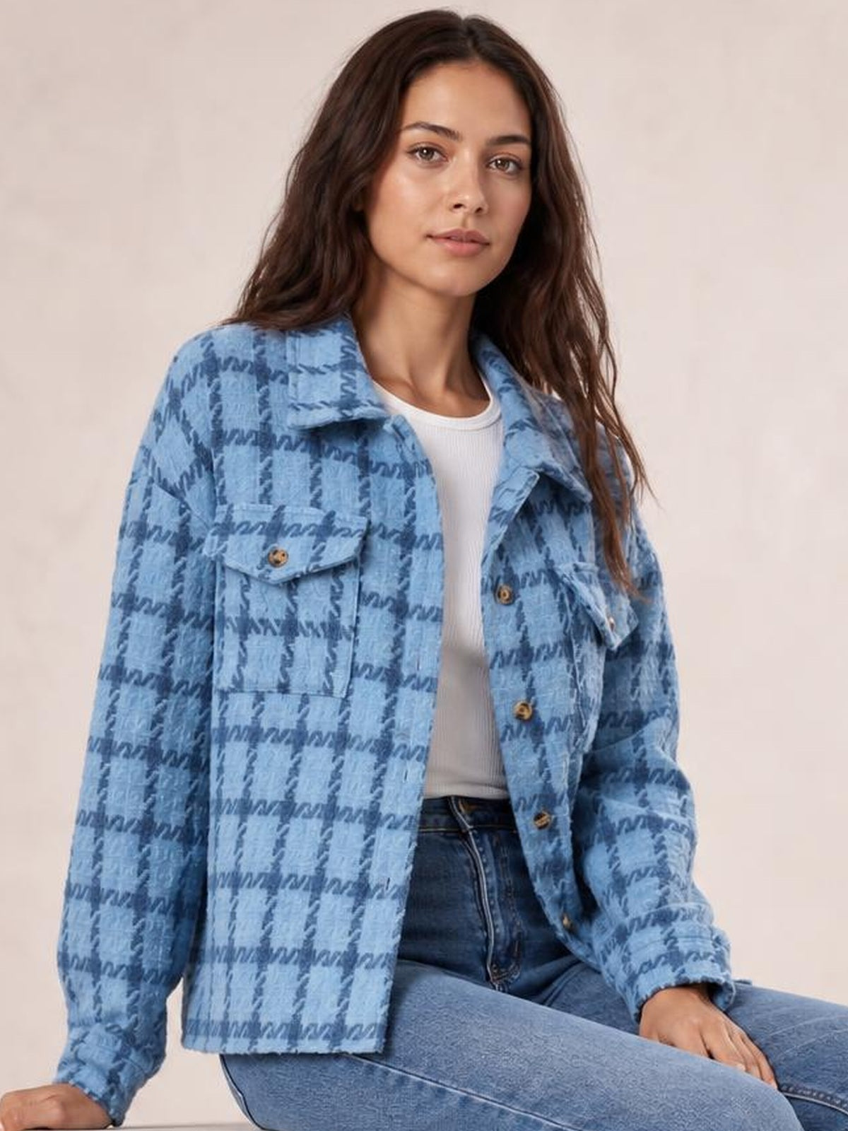 A seated model wears a light blue plaid button-up jacket with long sleeves, a pointed collar, flap chest pockets, and a relaxed straight silhouette.