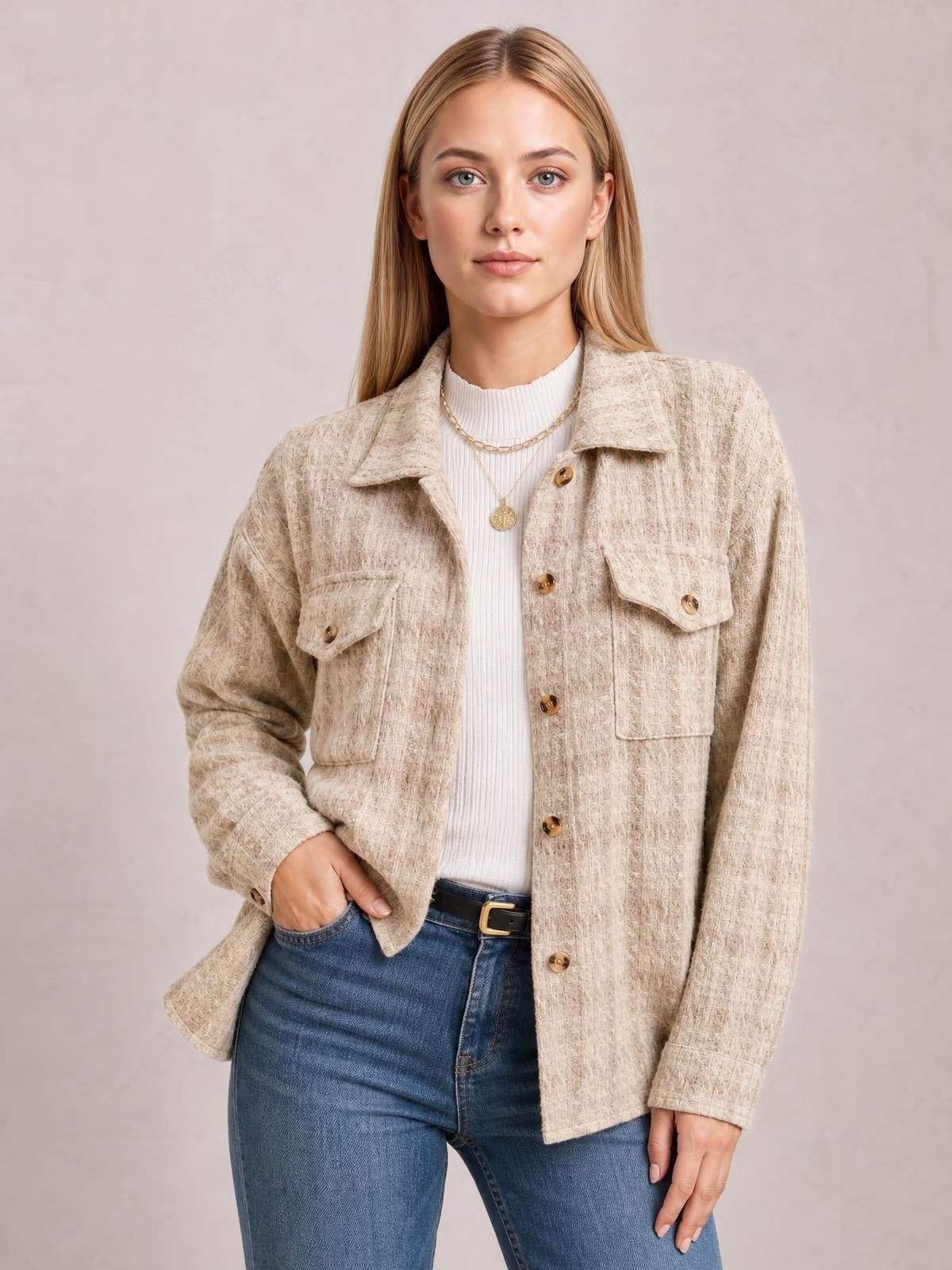 A model poses front-facing in a beige plaid button-up jacket with long sleeves, a collared relaxed silhouette, and flap pockets, one hand resting in her jeans pocket.