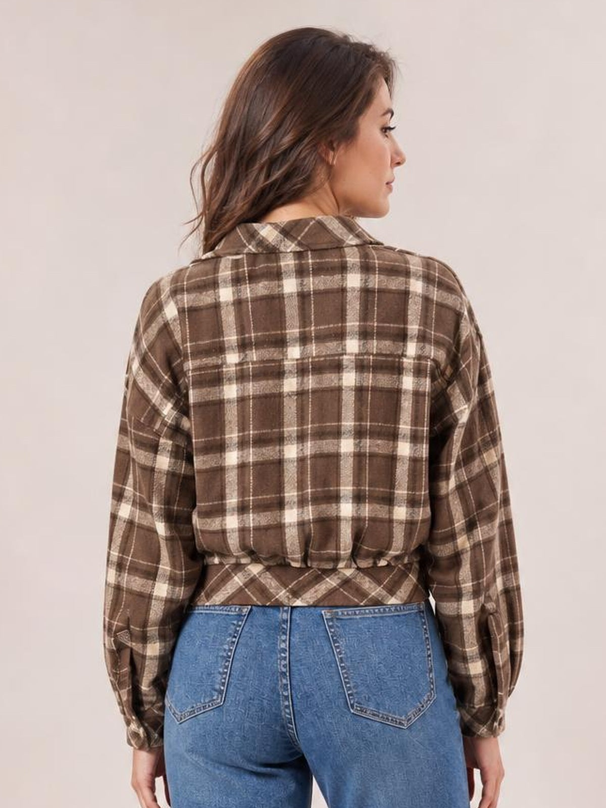 A woman poses with her back to the camera, showcasing a brown-and-cream plaid button-up jacket with a collar, long sleeves, and a waist-length silhouette.