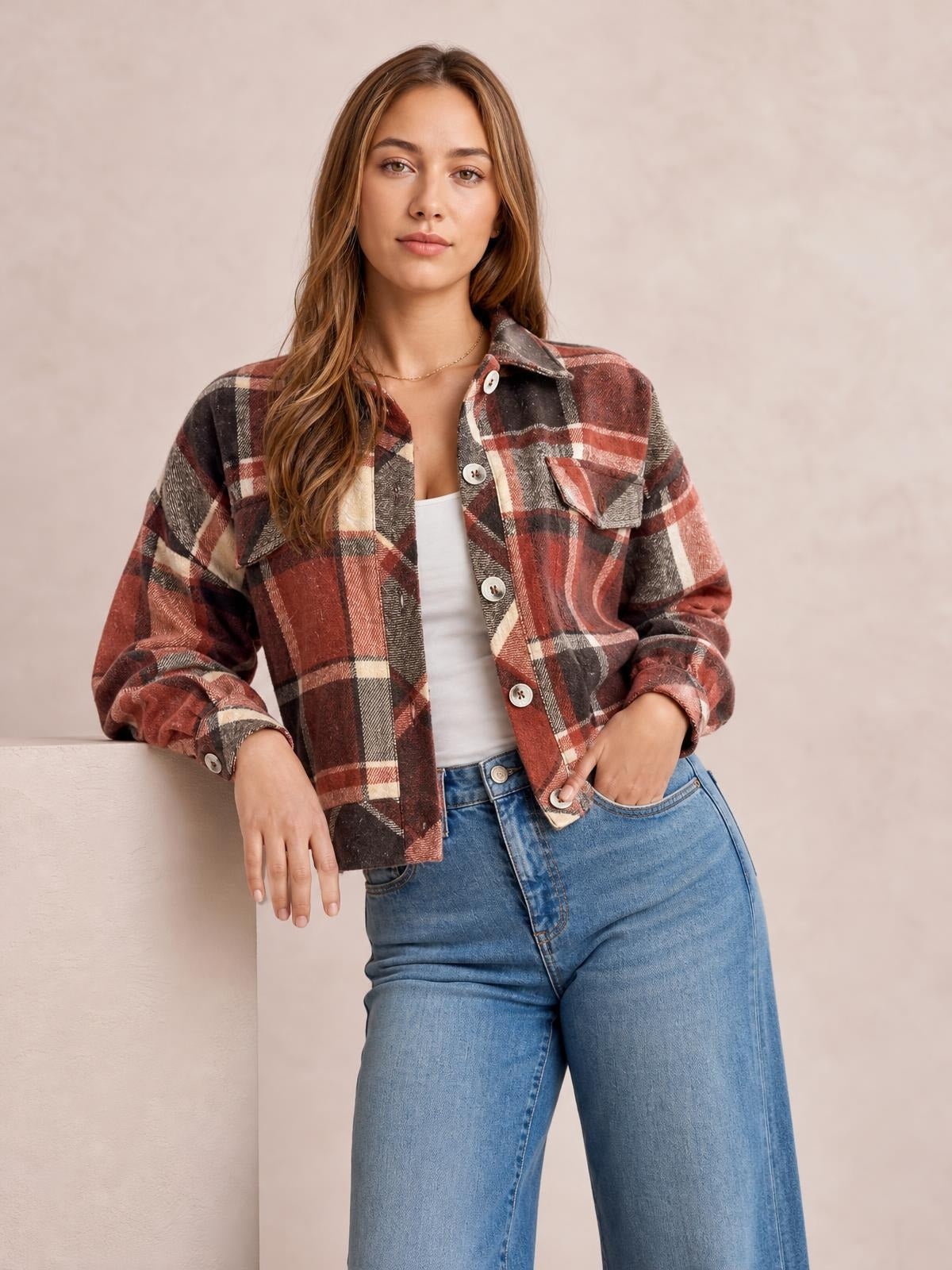 Model wearing a rust, black, and cream plaid button-up long sleeve jacket with a collared neckline, chest pockets, rolled cuffs, and a relaxed cropped silhouette.