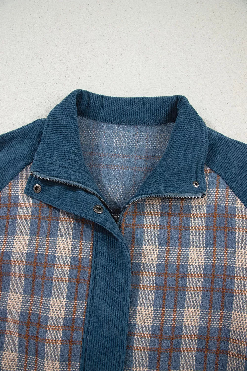Close-up of a blue plaid patchwork jacket with a corduroy collar, trim, front zipper, and snap-button closure.
