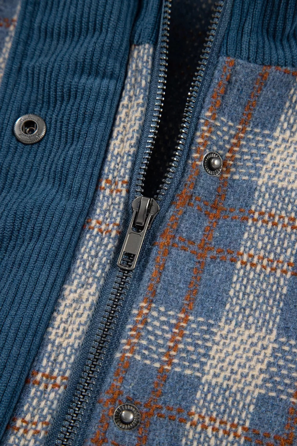 Close-up of a blue corduroy patchwork jacket showing a plaid panel, metal front zipper, and snap-button fastening.