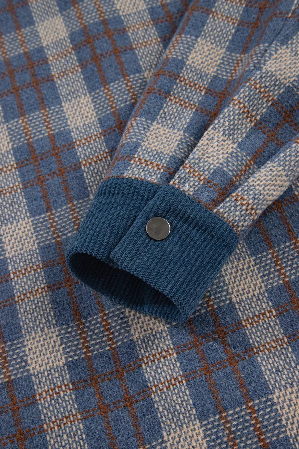 Close-up of a blue plaid patchwork jacket sleeve showing a ribbed corduroy cuff finished with a single snap button.