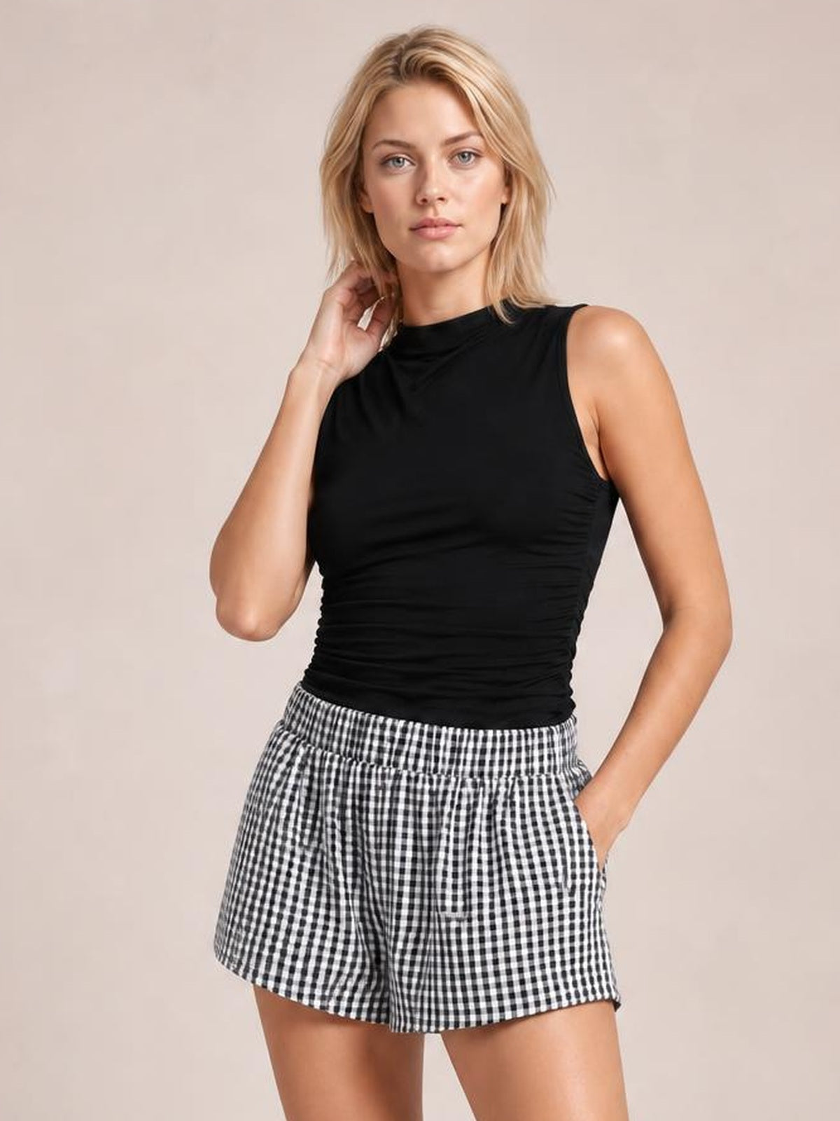 A woman modeling a black sleeveless ruched top and black-and-white gingham check shorts against a solid beige studio background.