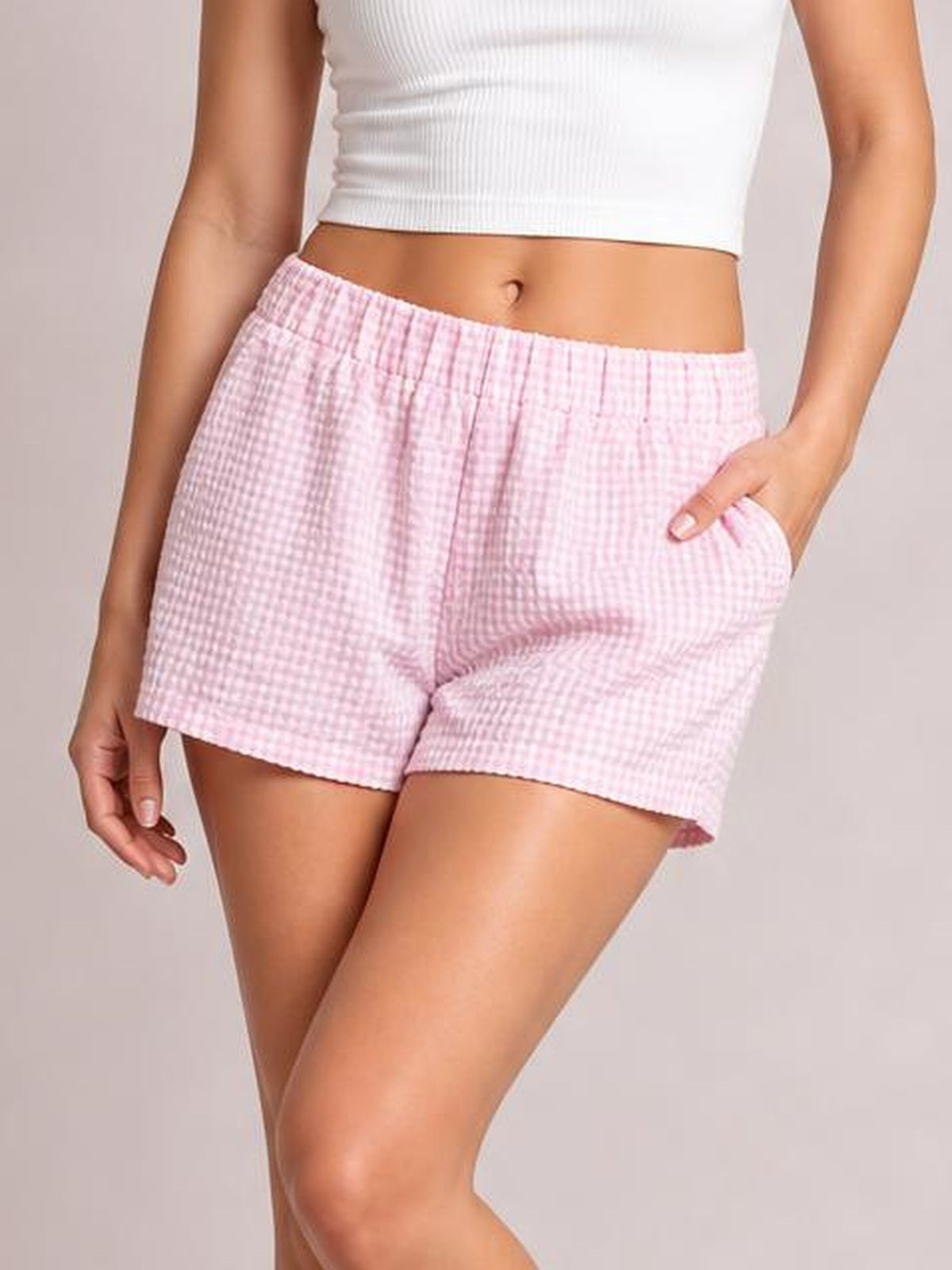 Model wears pink plaid elastic waist shorts with a relaxed, straight-leg silhouette, side pockets, and a hand resting in one pocket.