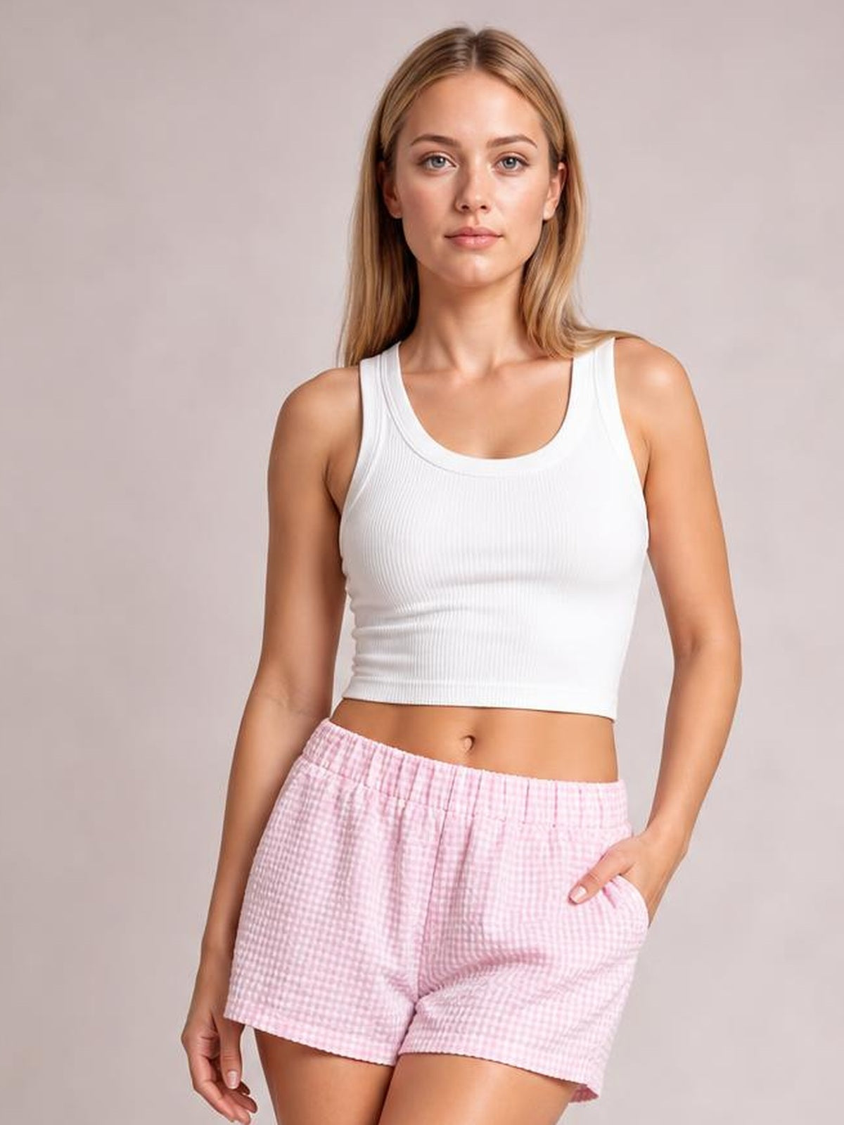 A woman modeling a white ribbed tank top and pink gingham shorts against a solid light gray studio background.