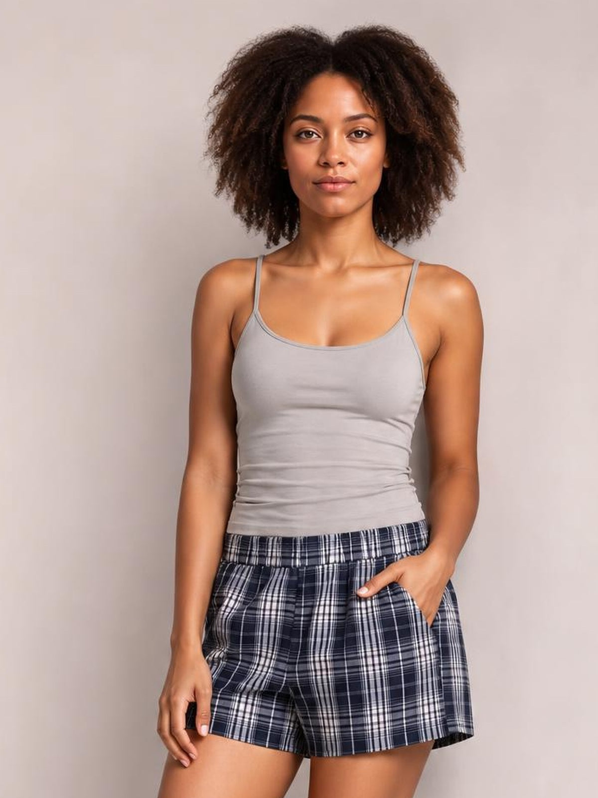 A woman modeling a light gray ribbed tank top and navy blue plaid shorts against a plain light gray studio background.