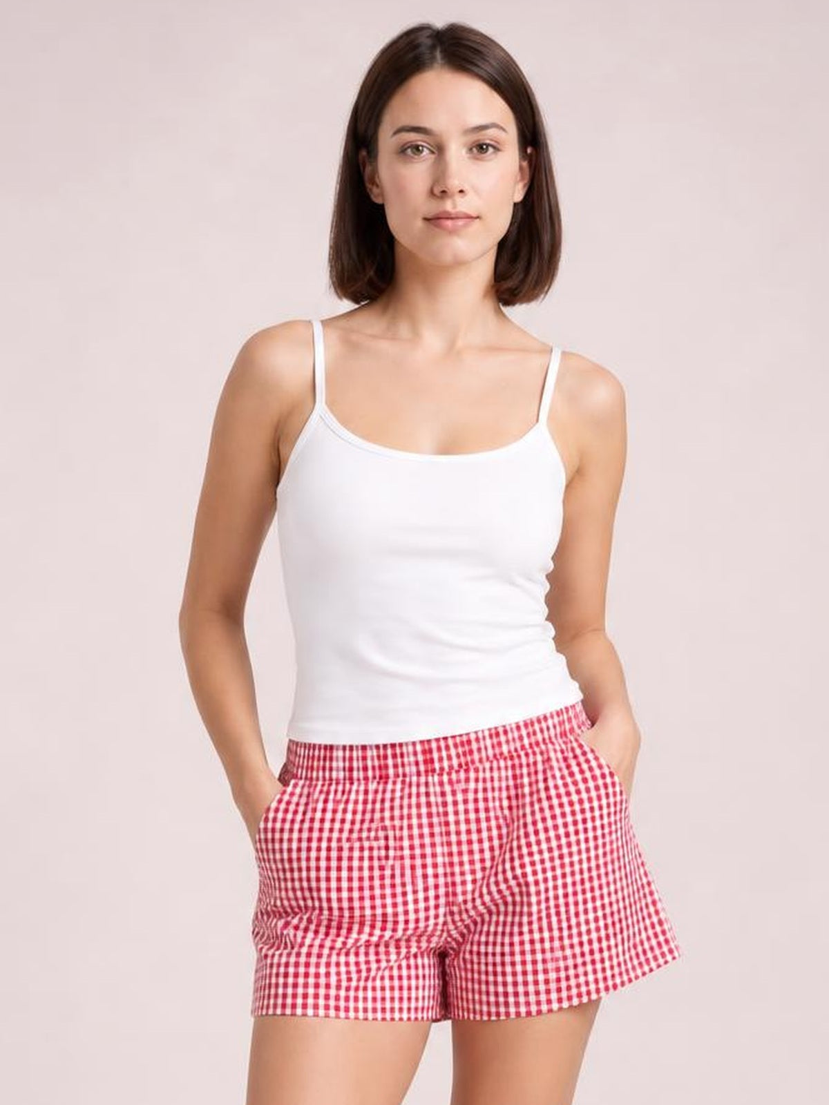 A woman modeling a white camisole tank top and red-and-white gingham check shorts against a solid pale pink studio background.