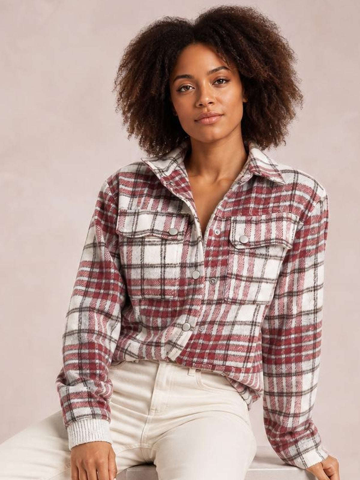 A seated model wears a red, white, and black plaid flannel shift jacket with a collar, long sleeves, metal buttons, and two chest pockets.