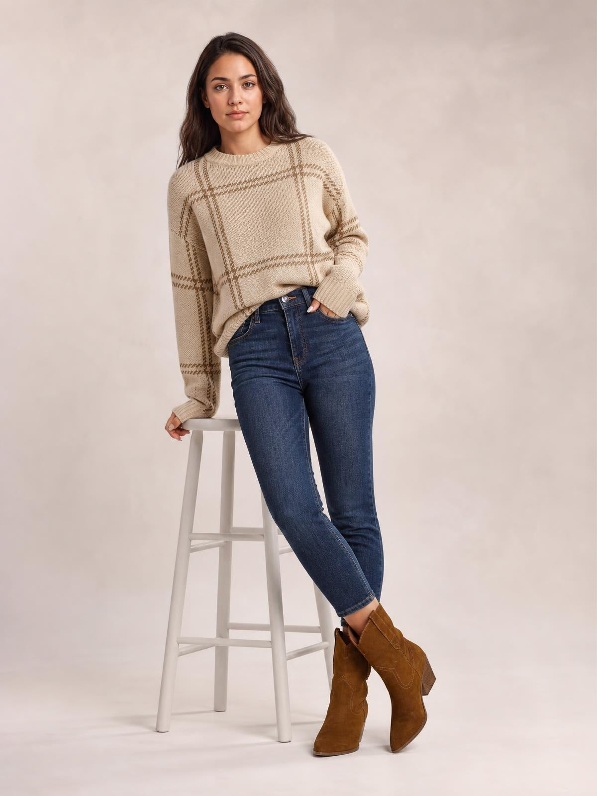 A model poses in a beige plaid round neck dropped shoulder sweater with a relaxed silhouette, ribbed neckline and cuffs, and one hand in her jeans pocket.