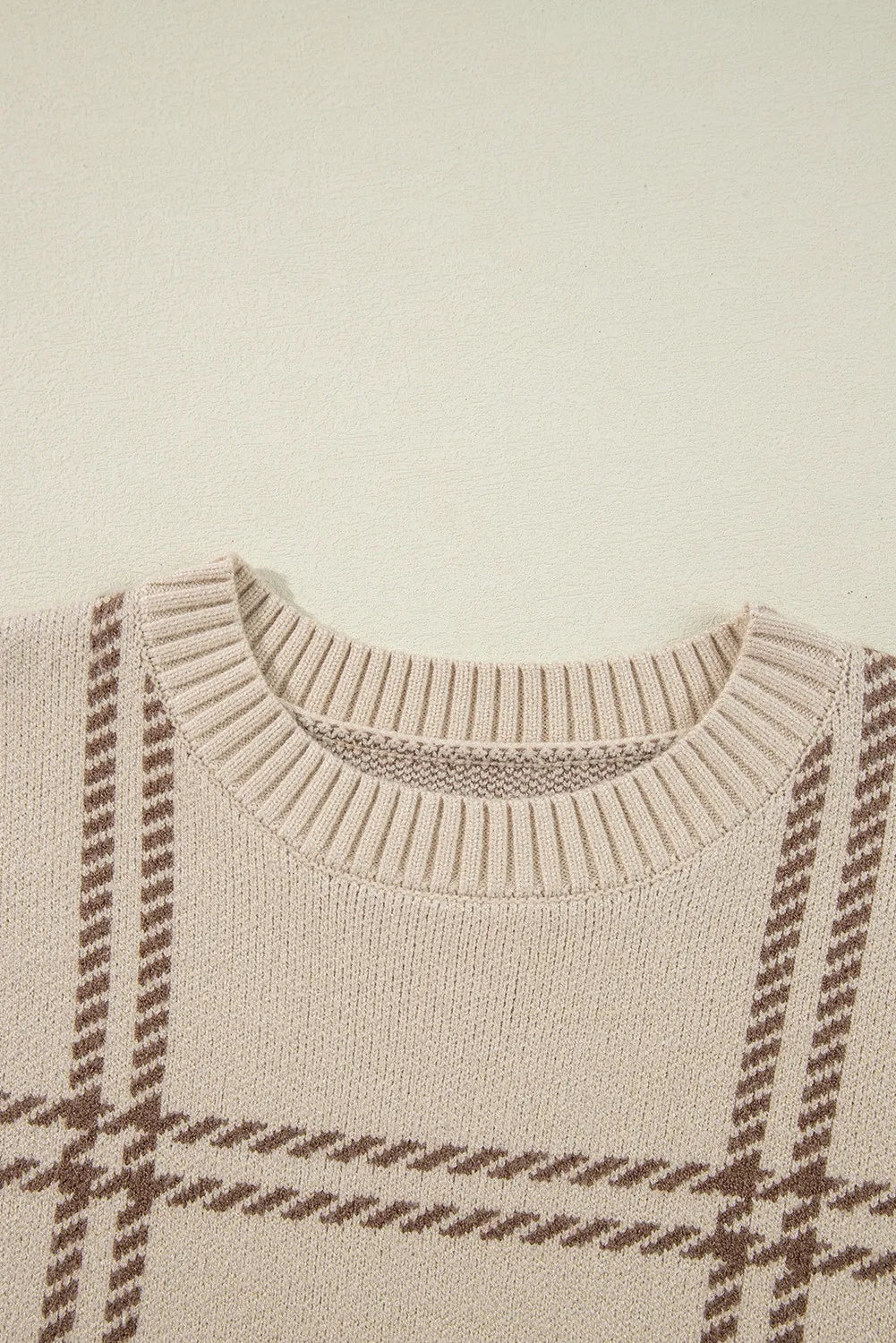 Close-up of a beige plaid sweater with brown patterning, a ribbed round neck, and textured knit construction.