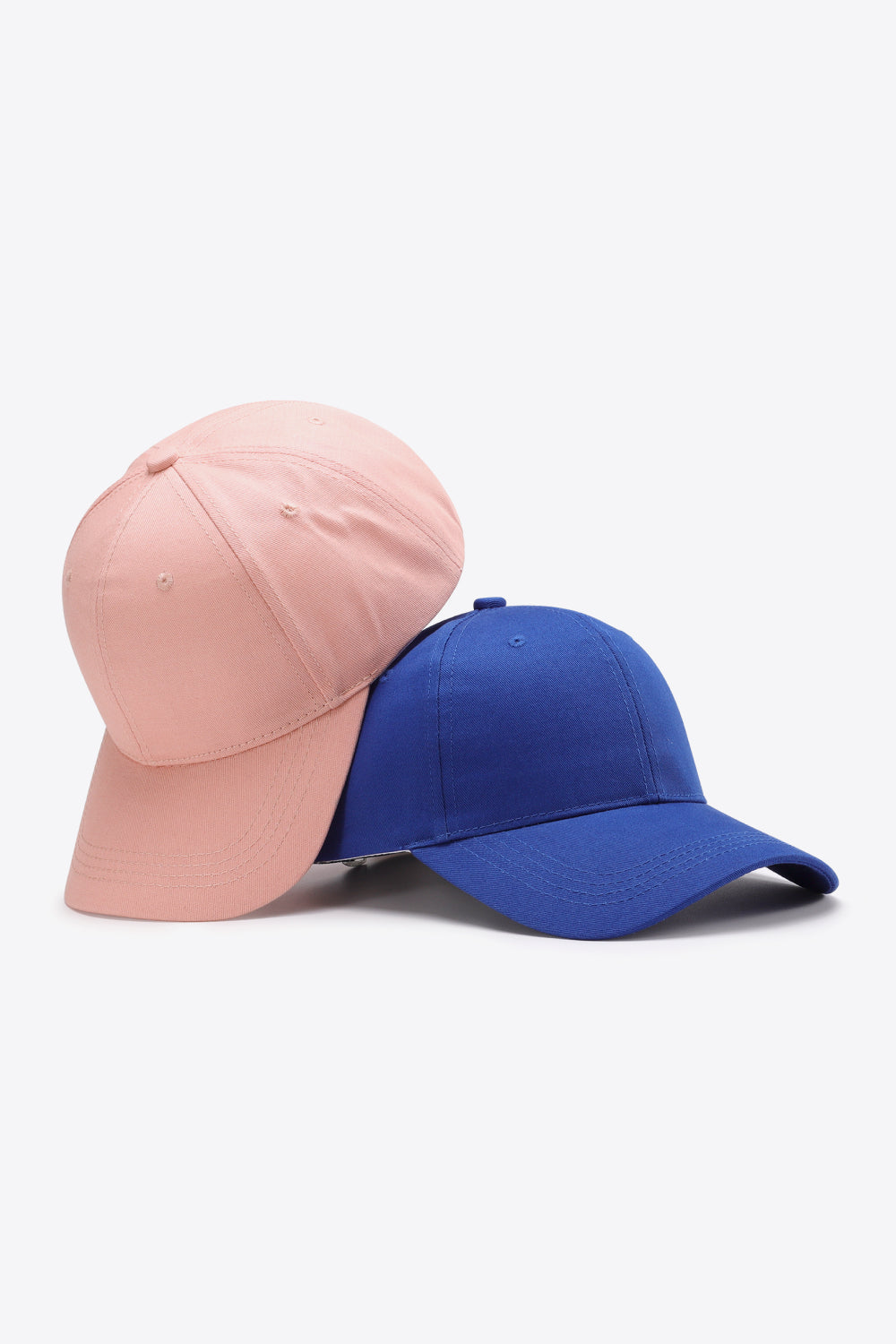 Two solid cotton baseball caps, one blush pink and one royal blue, feature structured six-panel crowns, curved brims, tonal stitching, and ventilation eyelets.