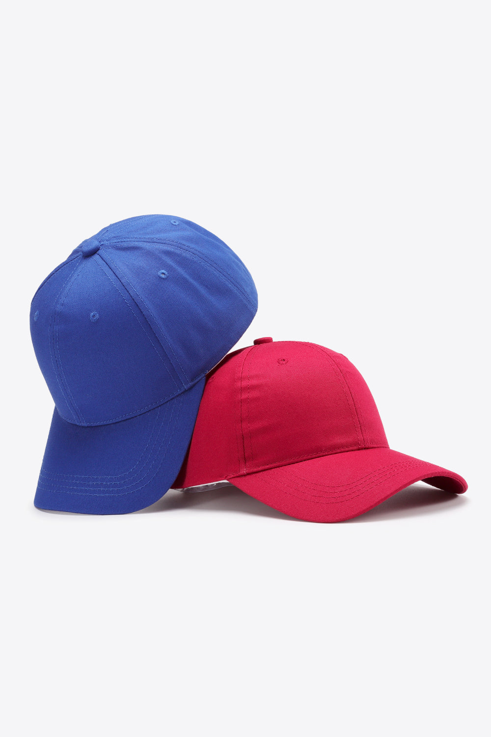 Two plain cotton baseball caps in royal blue and red feature structured six-panel crowns, tonal stitching, ventilation eyelets, and gently curved brims.