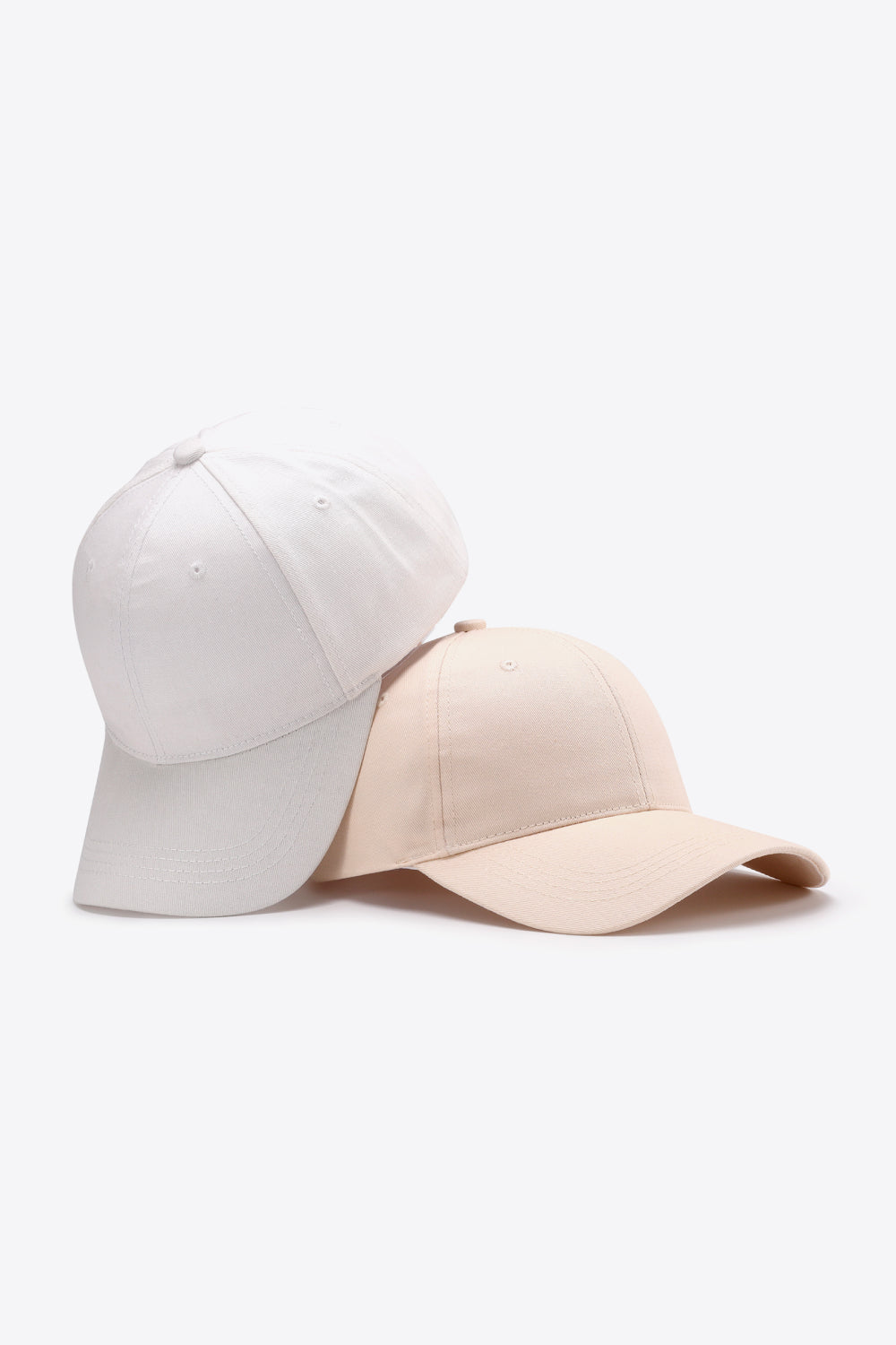Two solid cotton baseball caps in white and beige feature structured six-panel crowns, gently curved brims, tonal stitching, and ventilation eyelets.