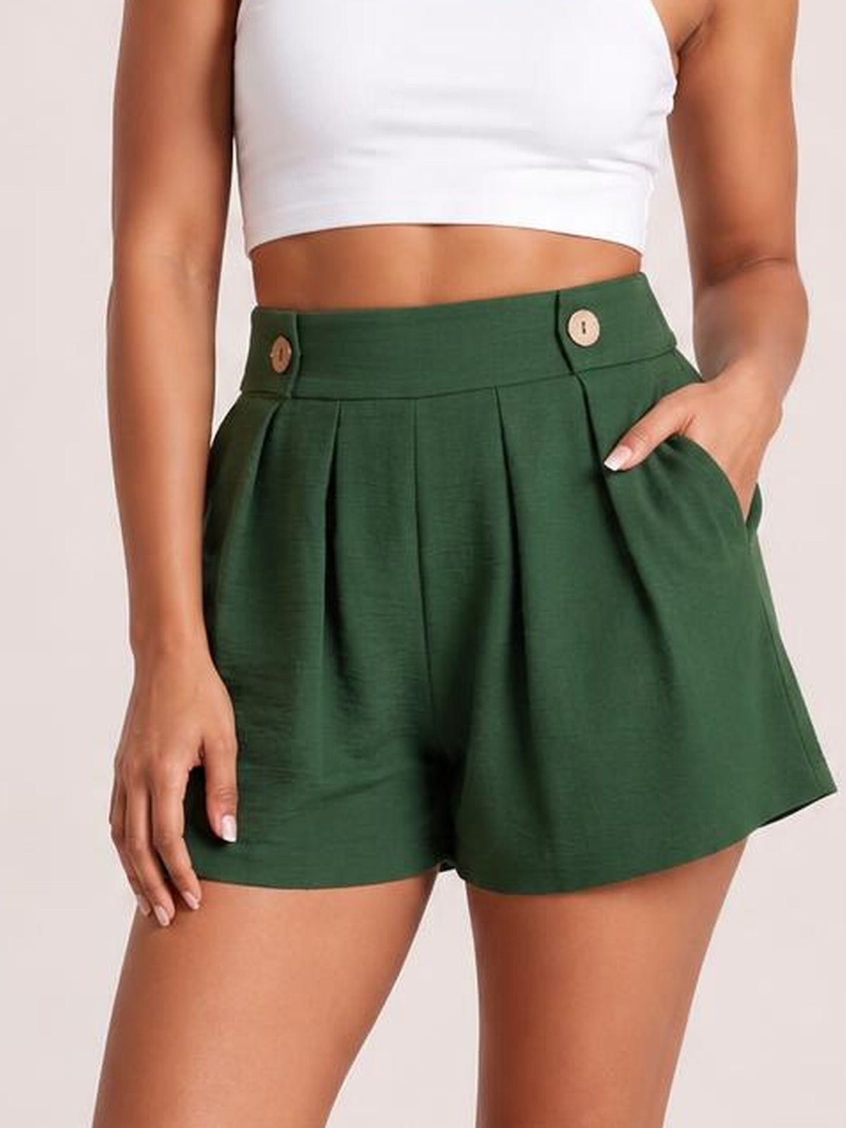 Model wears dark green pleated shorts with a high waistband, decorative buttons, roomy side pockets, and a relaxed wide-leg silhouette.