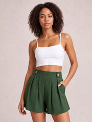 A woman modeling a white spaghetti-strap crop top and high-waisted forest green pleated shorts with gold-tone buttons, set against a plain beige studio background.