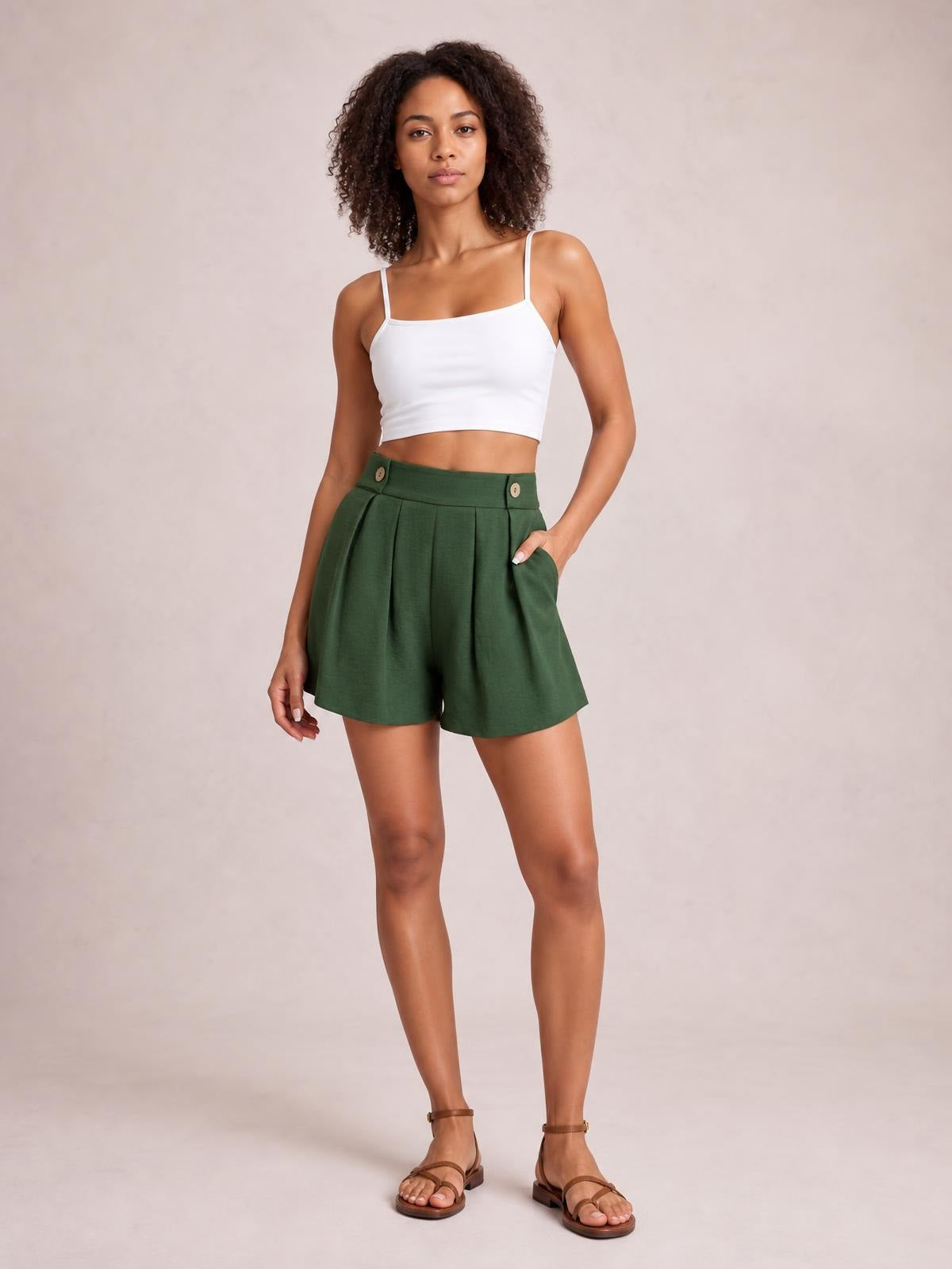 Model wears dark green pleated shorts with a high waistband, decorative buttons, side pockets, and a relaxed wide-leg silhouette, posing with one hand in a pocket.