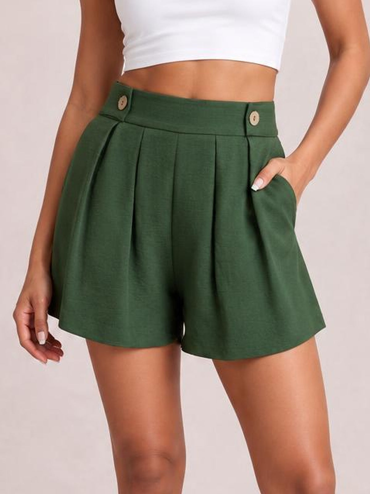 A model wears forest green pleated shorts with a high waistband, decorative buttons, side pockets, and a relaxed wide-leg silhouette, posing with one hand in a pocket.