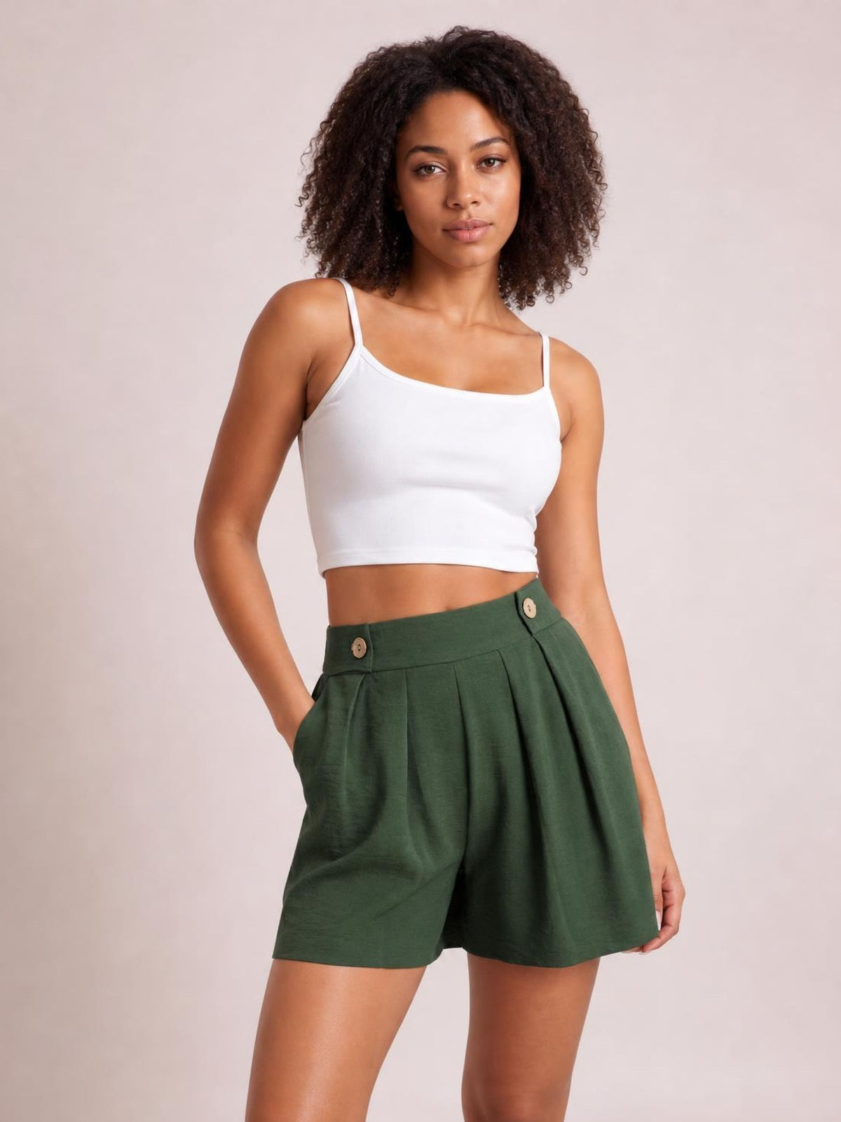 Model poses with one hand in the pocket of dark green pleated shorts featuring a wide waistband, decorative buttons, and a relaxed silhouette.