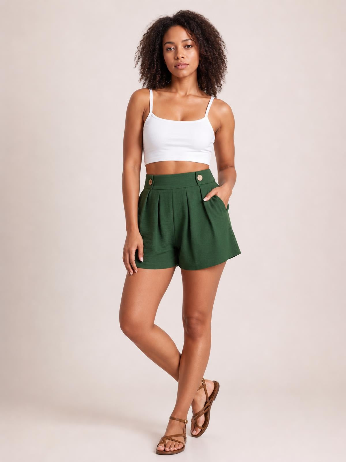 A model poses in forest green pleated shorts with a high waistband, decorative buttons, side pockets, and a relaxed wide-leg silhouette.