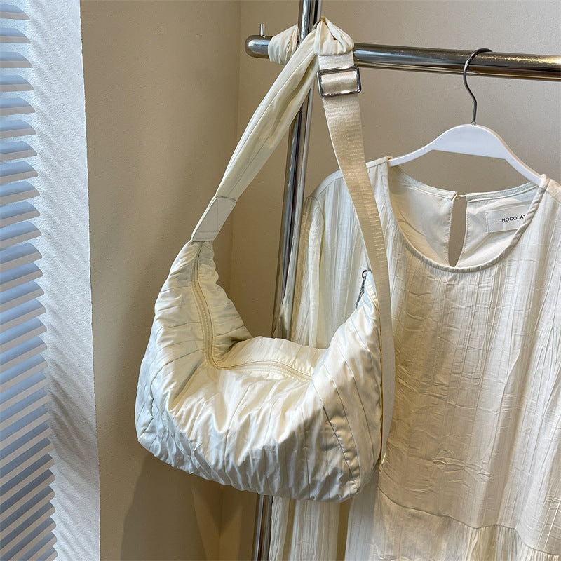 A cream Pleated Dumpling Crossbody Bag features a softly sculpted crescent silhouette, gathered pleats, and a broad adjustable shoulder strap.