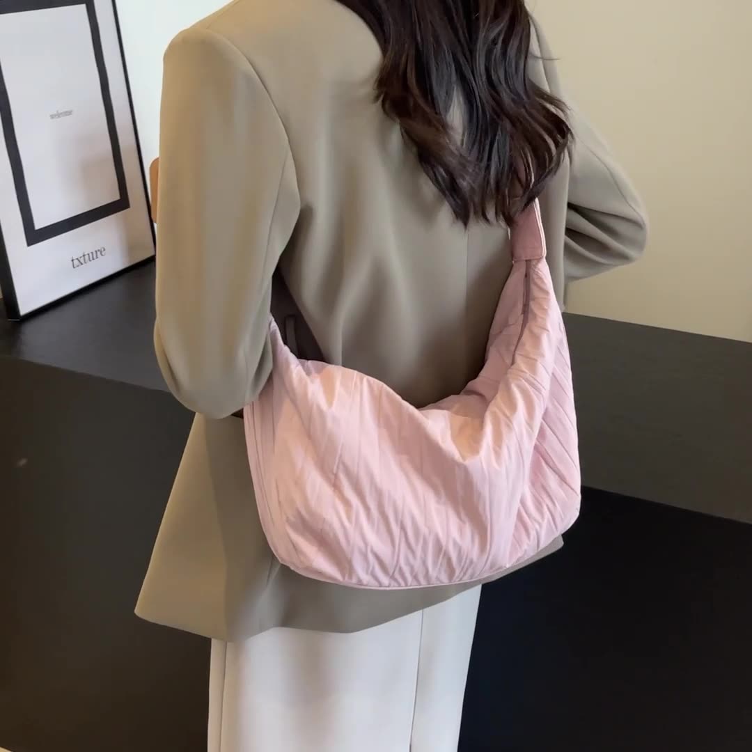 A model wears the pink Pleated Dumpling Crossbody Bag over one shoulder, showing its soft crescent silhouette, gathered pleats, slouchy shape, and broad strap.