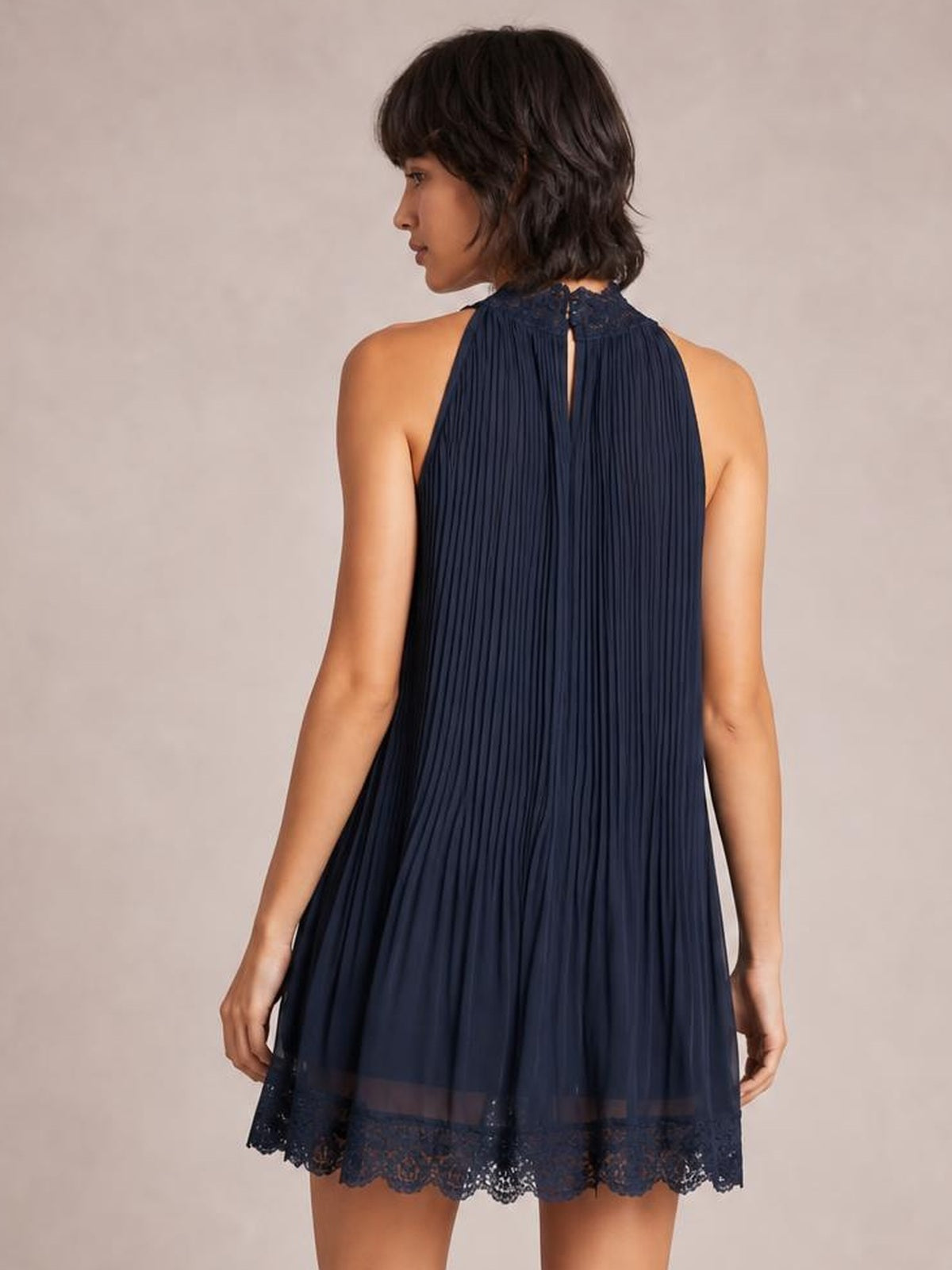 A model looks over her shoulder in a navy sleeveless mini dress with a pleated, swing silhouette, lace-trimmed neckline and hem, and a back cutout.