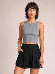 A woman modeling a gray ribbed sleeveless crop top with a side cutout and a black pleated tennis skirt, set against a solid beige studio background.