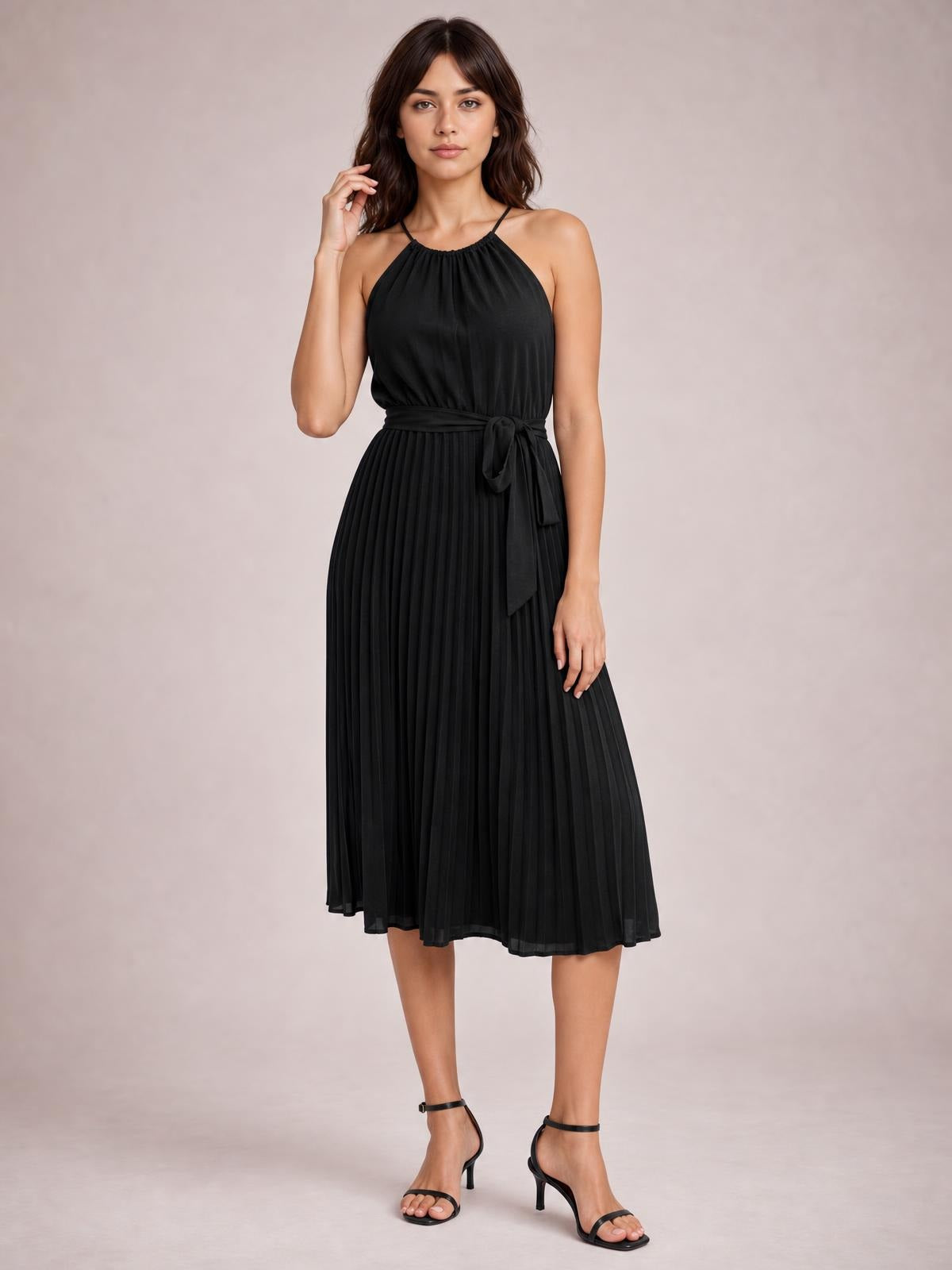 Model poses in a black pleated midi dress with spaghetti straps, a gathered neckline, tied waist, and flowing A-line silhouette.