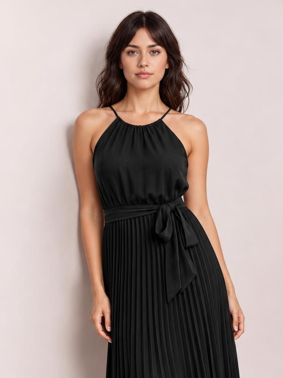 Model poses in a black pleated midi dress with spaghetti straps, a softly gathered bodice, and a tied waist creating a flowing A-line silhouette.