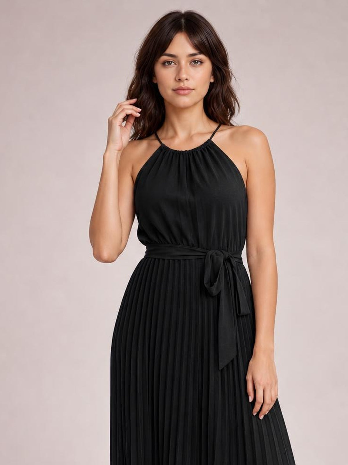 A model poses in a black pleated spaghetti strap midi dress with a gathered neckline, flowing silhouette, and tied waist sash.