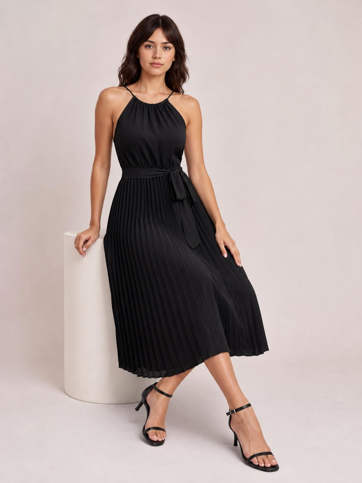 Model wearing a black pleated spaghetti strap midi dress with a gathered neckline, defined waist, tied sash, and flowing A-line silhouette.