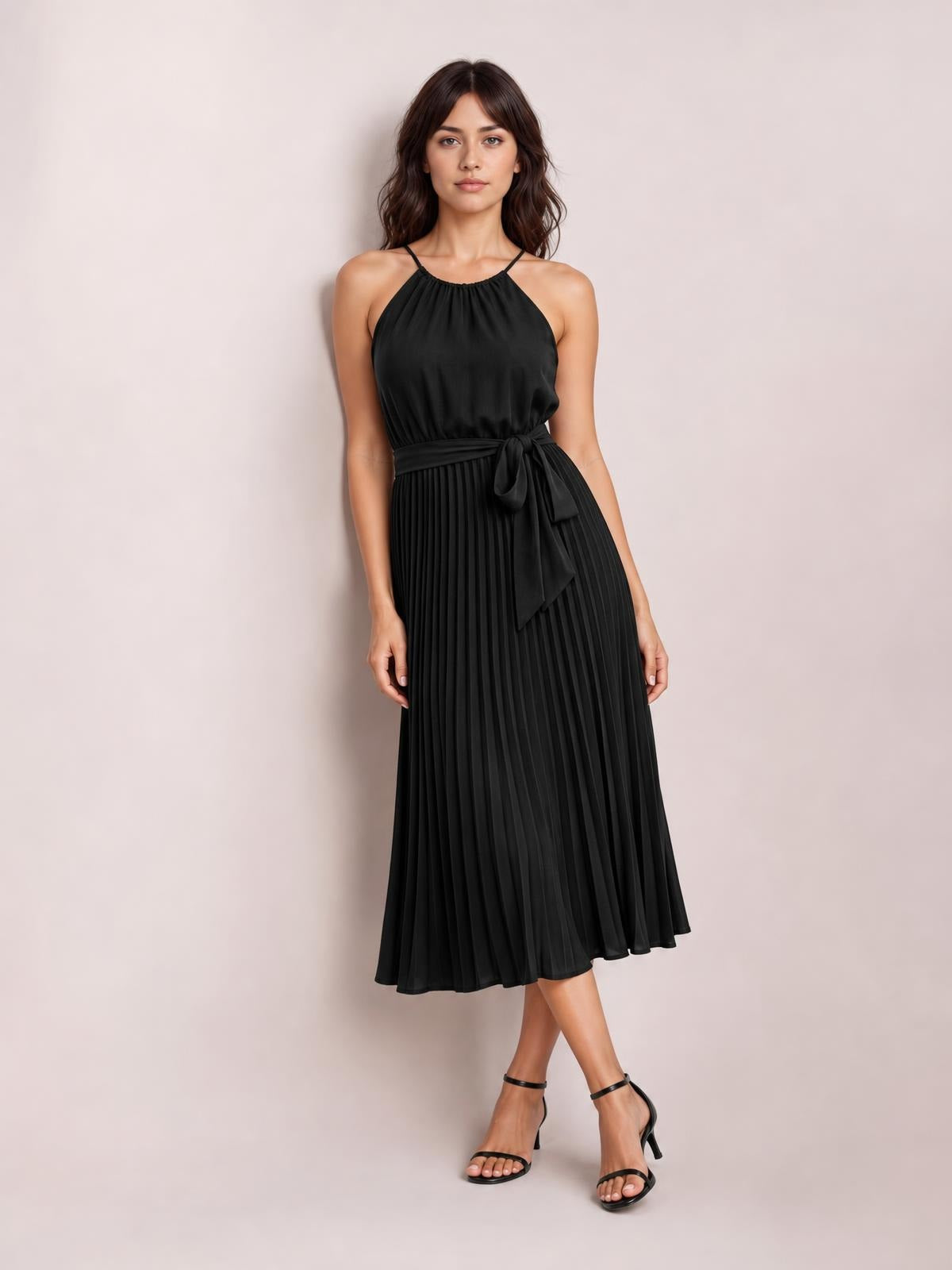 Model poses in a black pleated spaghetti strap midi dress with a tied waist, flowing A-line silhouette, and ankle-length hem.
