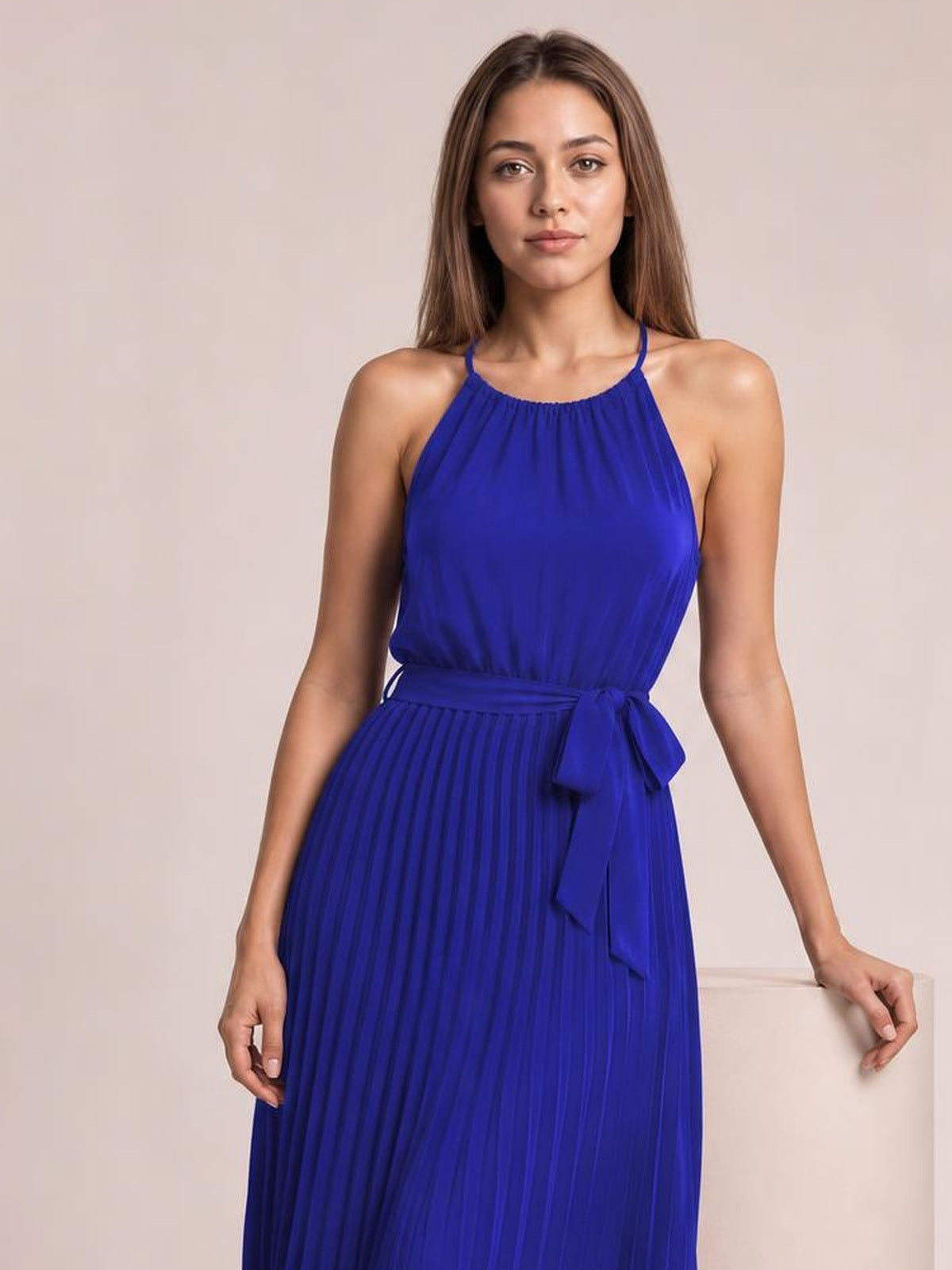 Model wearing a royal blue pleated midi dress with spaghetti straps, a gathered neckline, fitted waist, and tied sash, standing with one hand resting on a ledge.