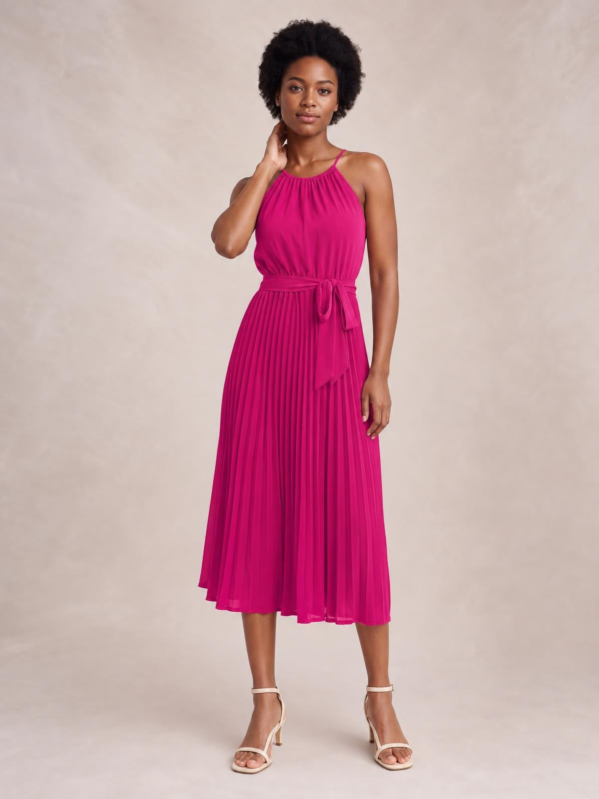 Model poses in a bright pink pleated spaghetti strap midi dress with a gathered neckline, defined waist, tie belt, and flowing A-line silhouette.
