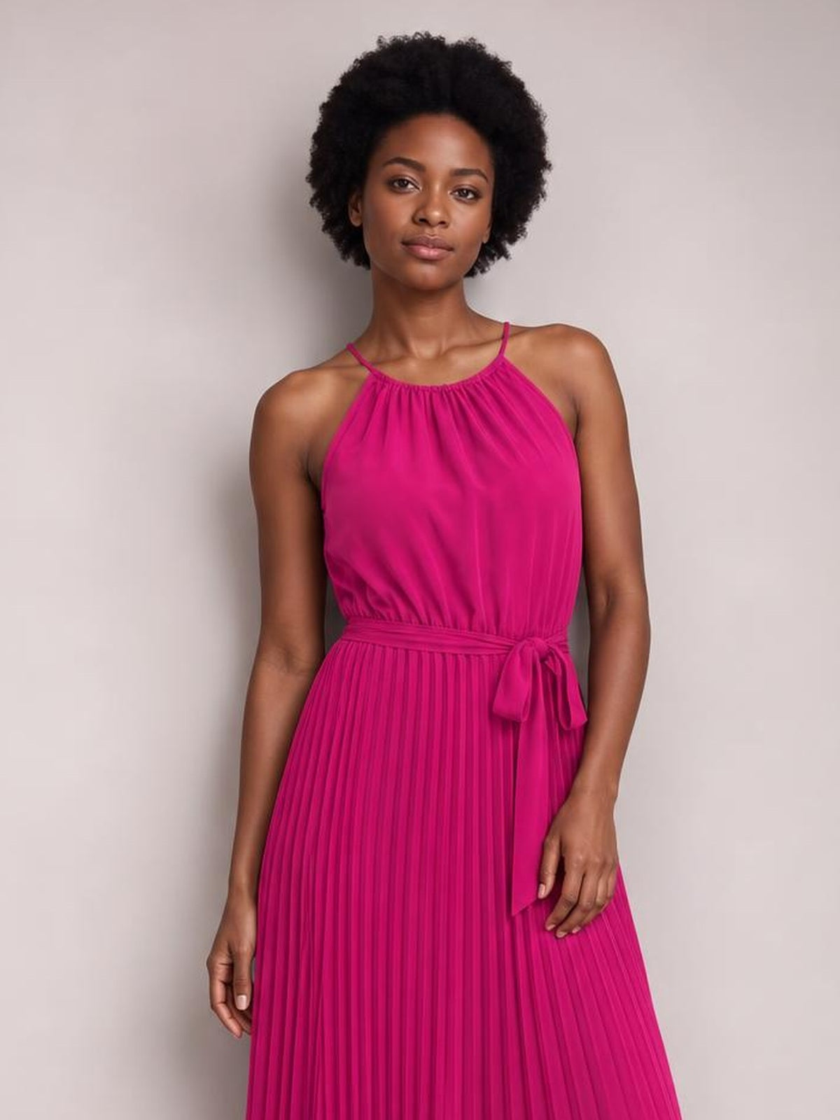 Model wears a bright pink pleated spaghetti strap midi dress with a gathered neckline, defined waist, tie belt, and flowing A-line silhouette.